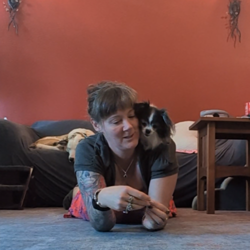 A woman lying on the floor with a small dog, both looking at a treat she is holding.