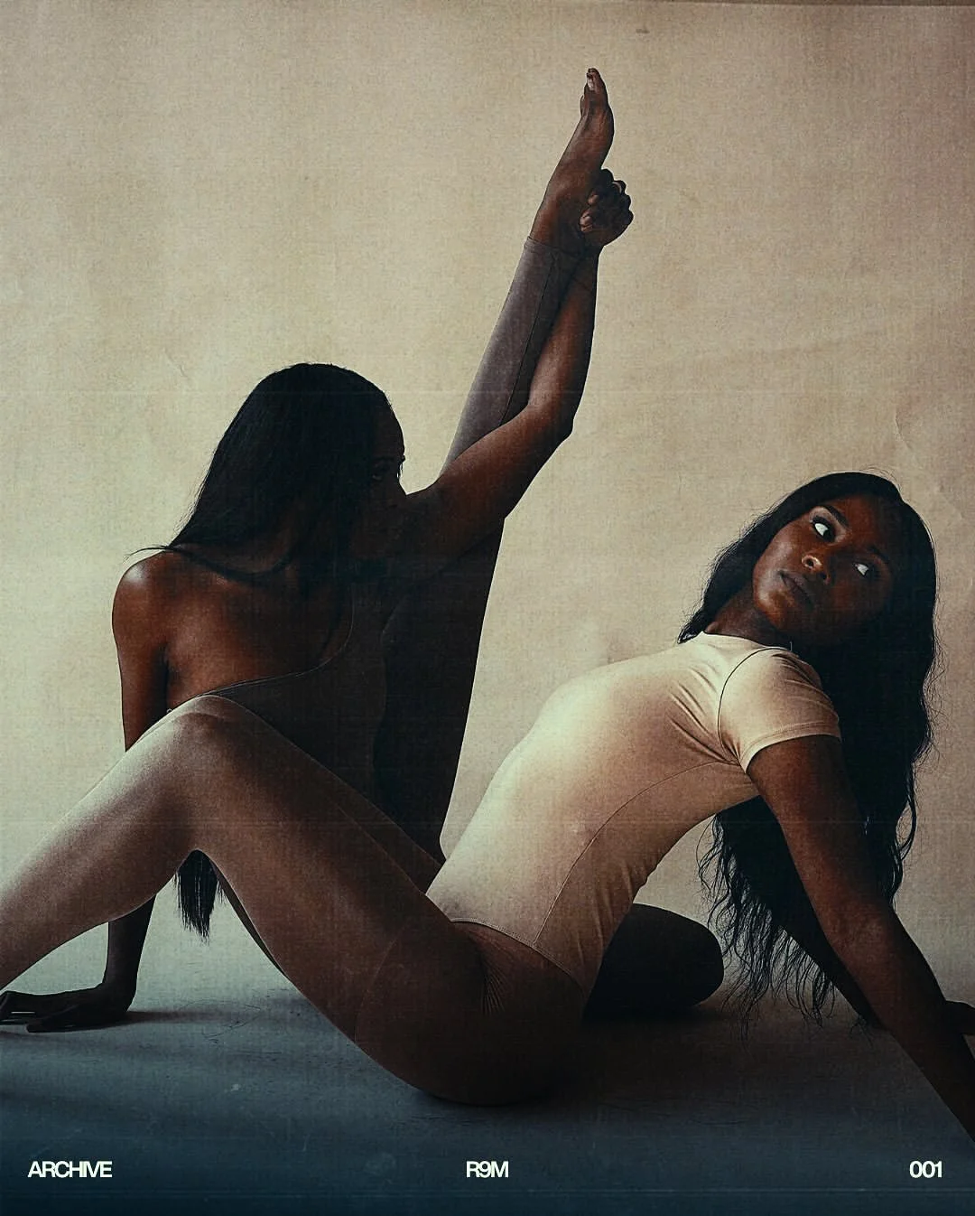 Two women in minimal clothing posing against a plain background, one sitting on the floor with legs bent and one stretching upward with her arm, both with dark hair.