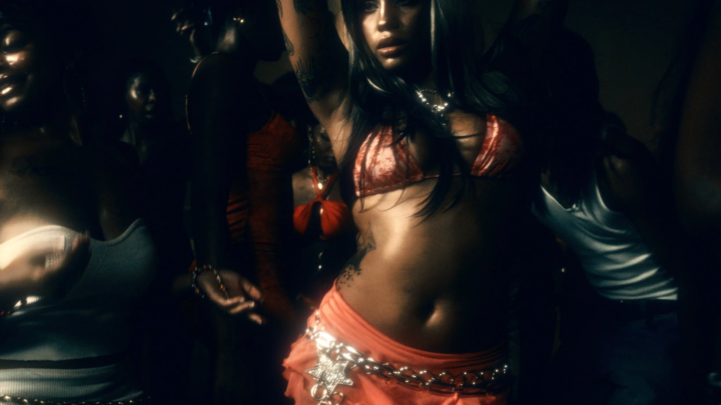 A woman with long black hair wearing a red velvet bra and orange pants with a star belt, surrounded by people in dark clothing in a dark environment.