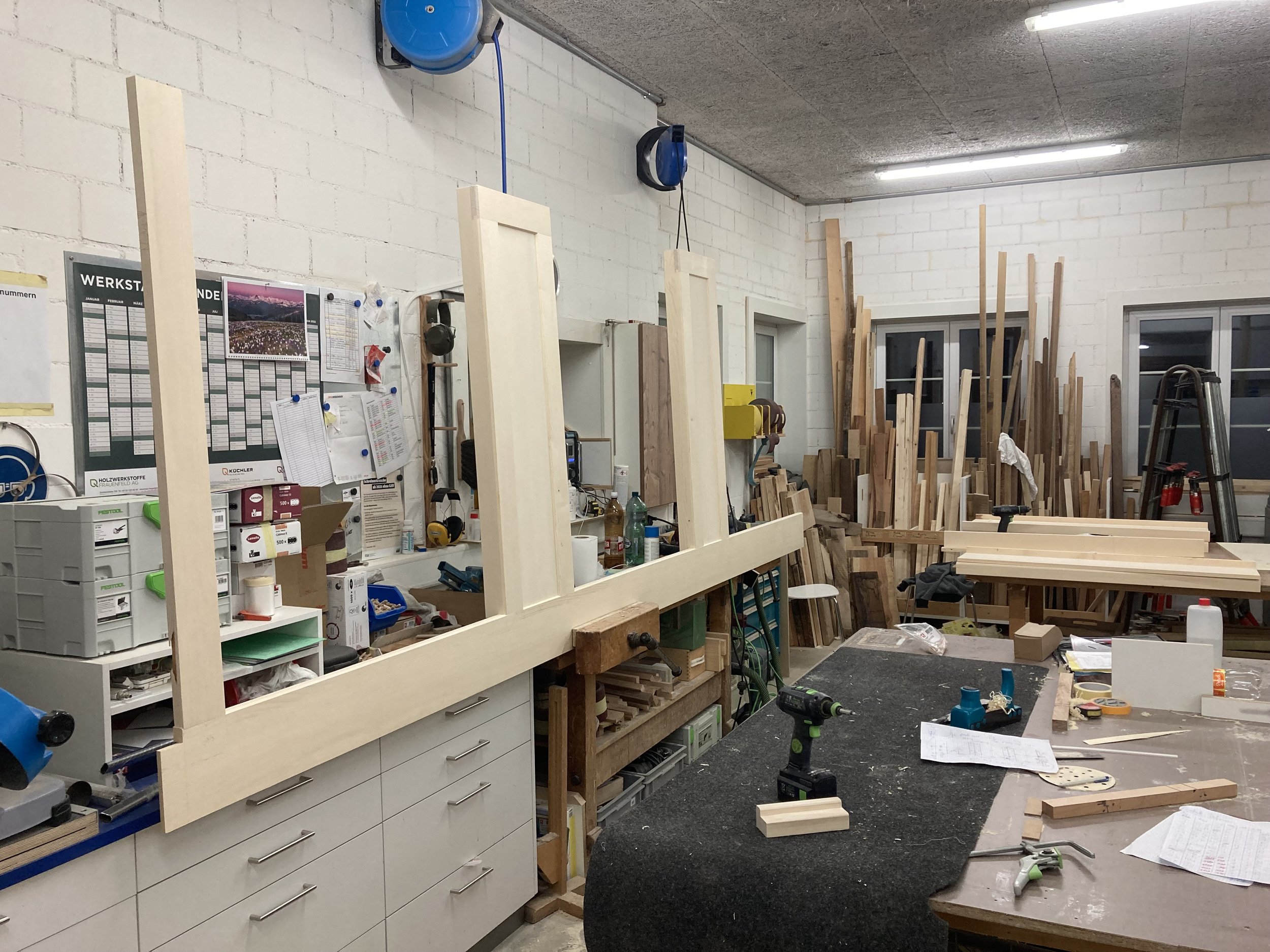 Werkstatt mit Holzbrettern, Werkzeugen und Materialien auf Arbeitsflächen; im Hintergrund stehen gestapelte Holzstücke.