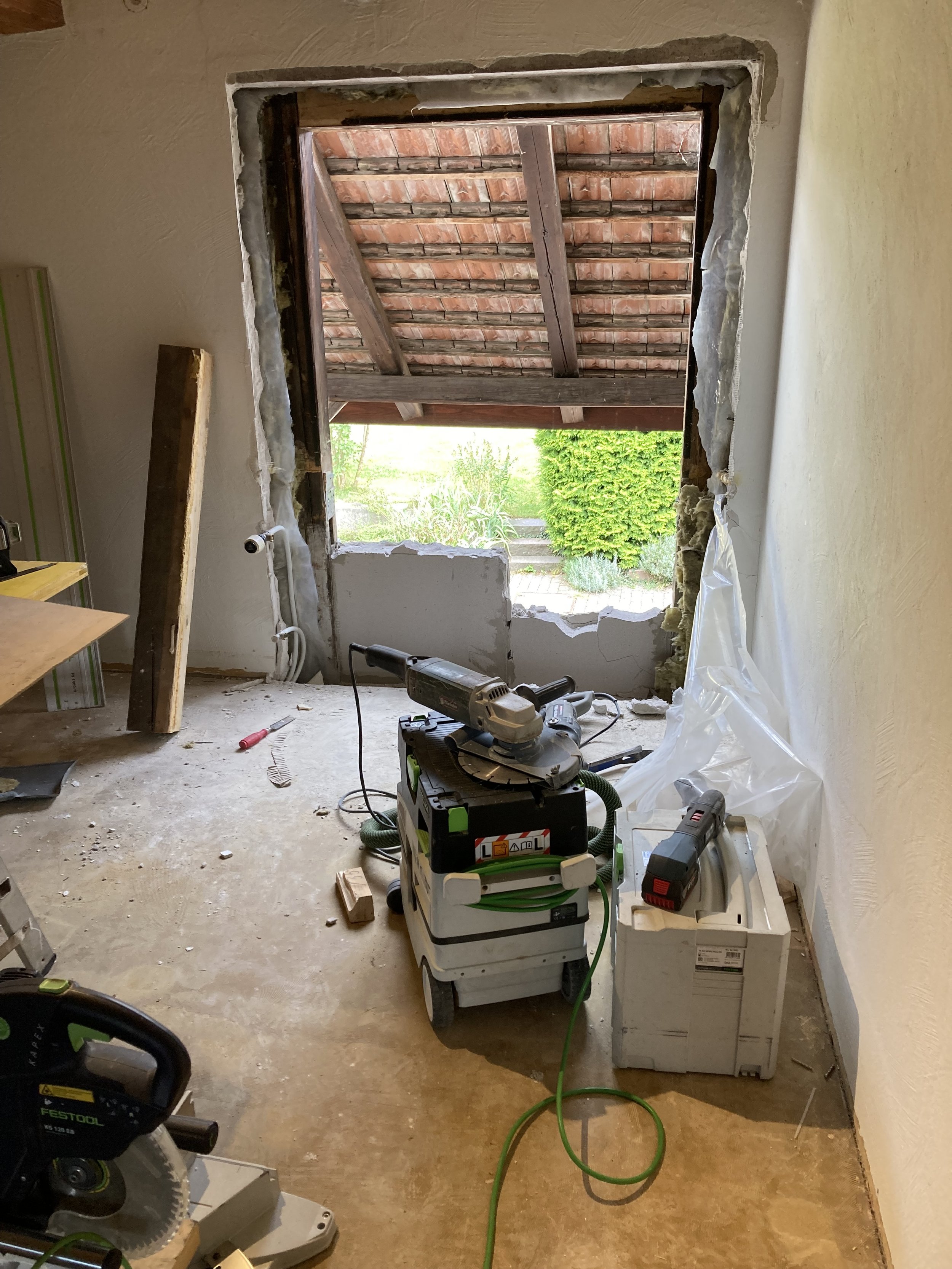 Innenraumszene mit Renovierungsarbeiten. Eine Wandöffnung zeigt ein freigelegtes Fenster oder Türrahmen. Werkzeuge wie eine Kreissäge, eine Schleifmaschine und andere Renovierungsgeräte sind auf dem Boden verteilt. Der Blick aus der Öffnung zeigt ein