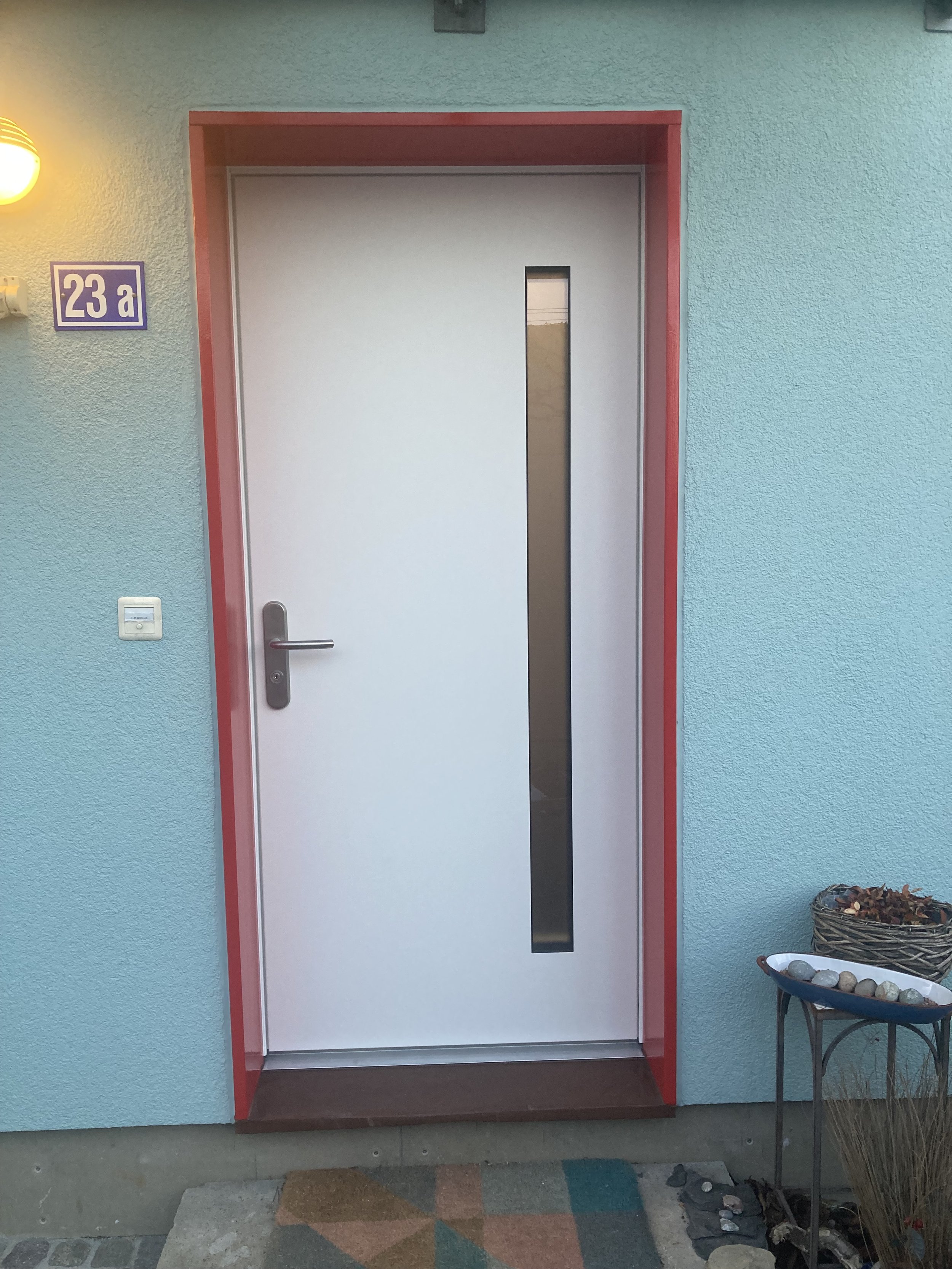 Weiße Tür mit rotem Rahmen und schmalem vertikalem Fenster, beige Wand, Hausnummer 23a. Links ist eine Lampe, rechts steht ein Pflanzentopf und ein Steintablett auf einem Tisch.