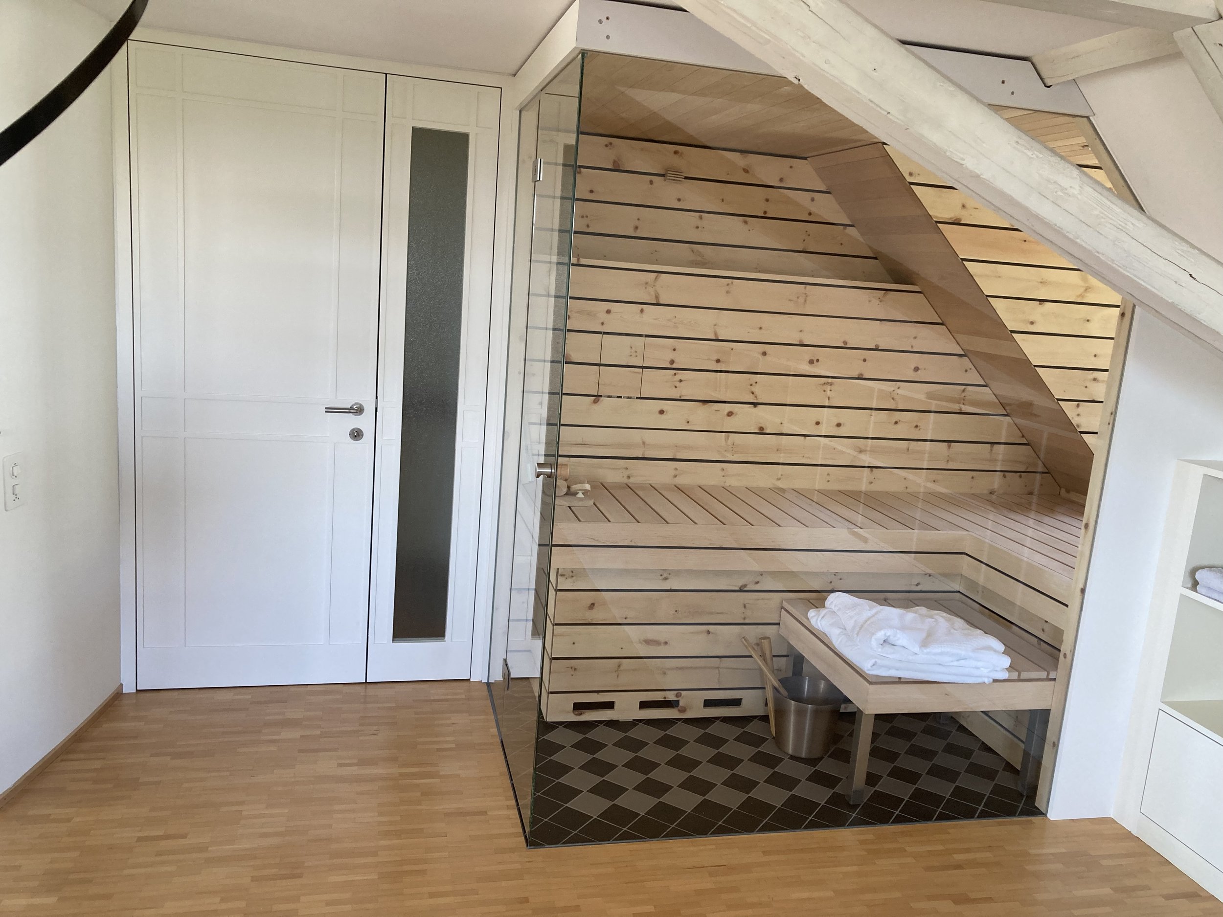 Moderne Sauna mit Holzverkleidung in einem Raum mit Parkettboden, Glastür und Handtüchern auf einem Holzregal.