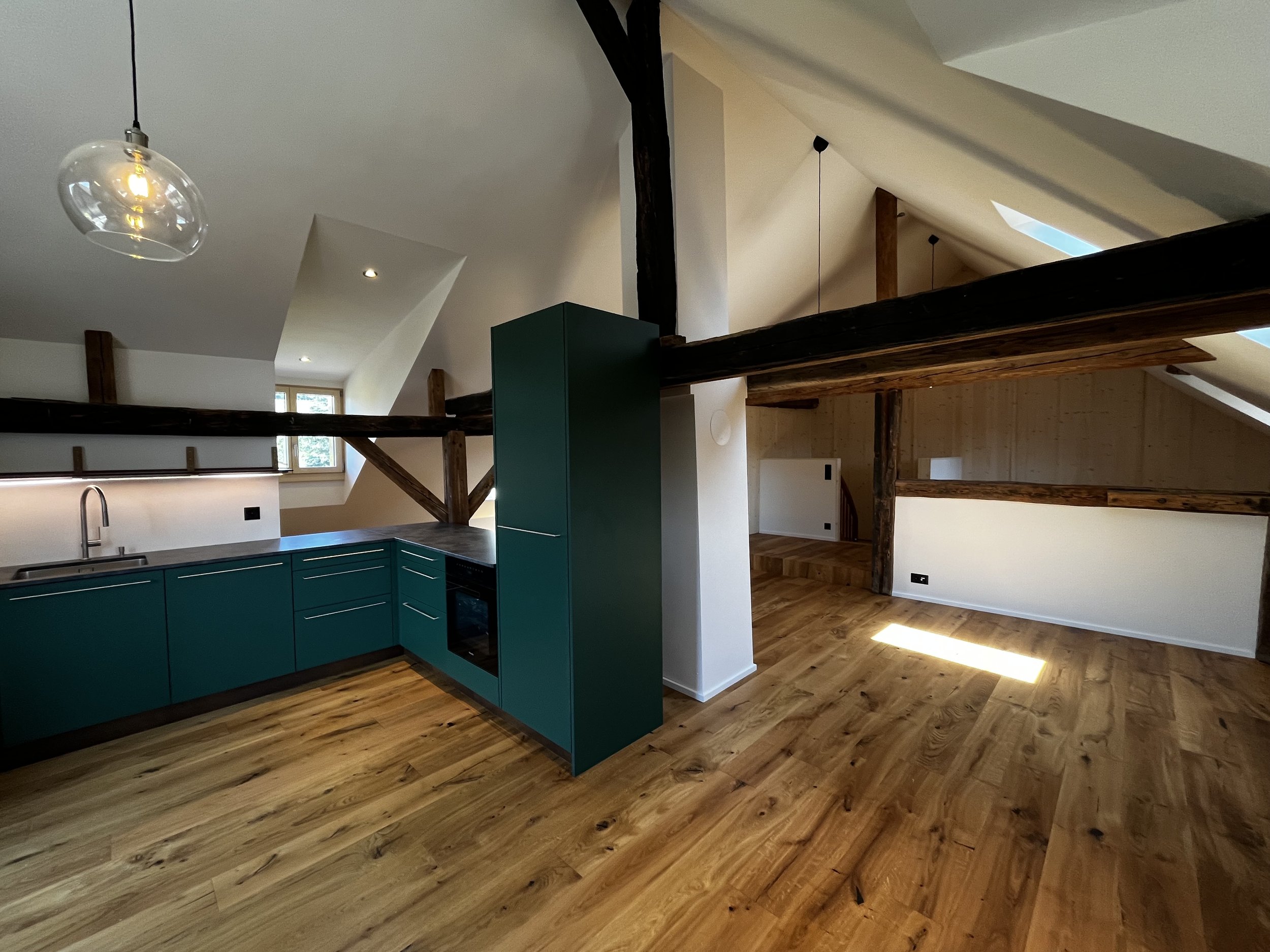 Renovierte Dachgeschosswohnung mit Holzbalken, modernen grünen Küchenzeilen und Holzfußboden. Große Fenster und Deckenleuchte sorgen für natürliche Beleuchtung.