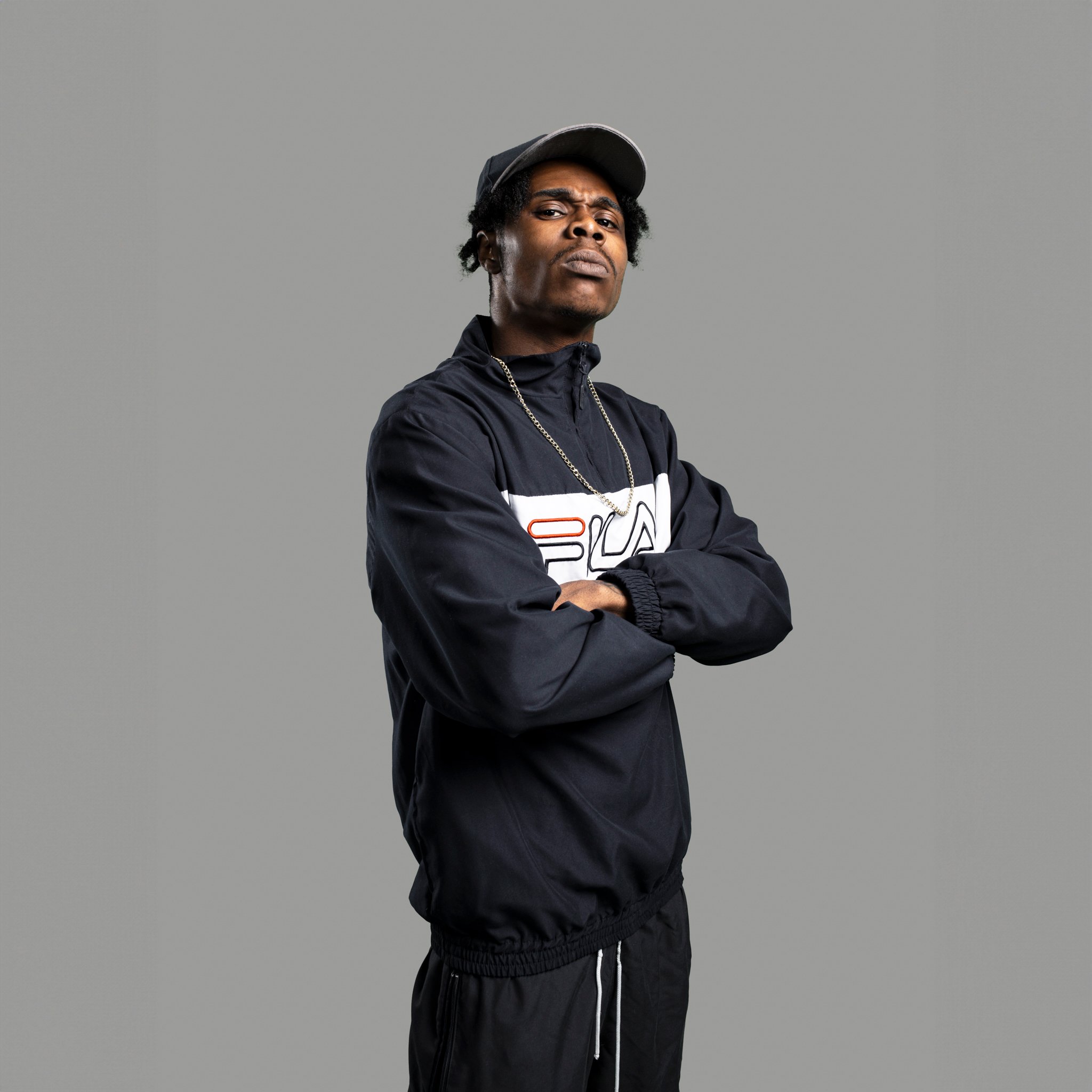 A young man with dark curly hair and a serious expression, wearing a black cap, black sports jacket with a white logo, and a gold chain, standing with arms crossed against a plain gray background.
