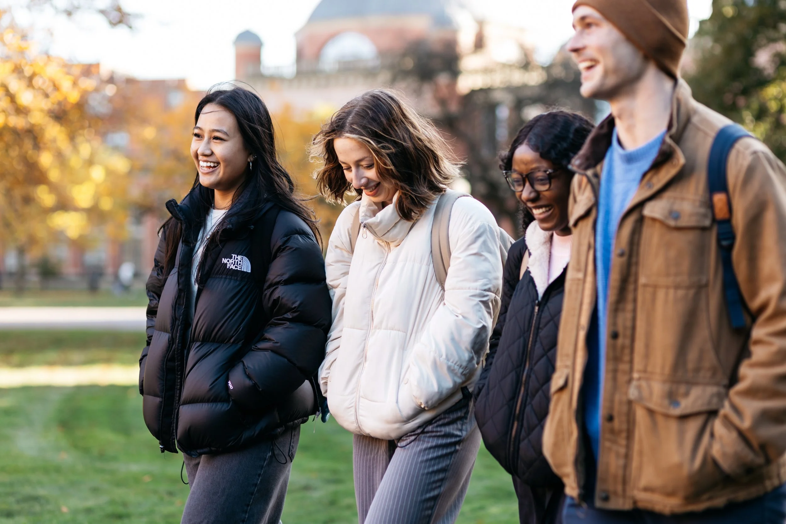 Group of four diverse friends walking outdoors on a fall day, smiling and laughing weather in jackets and backpacks, with trees and a building in the background.