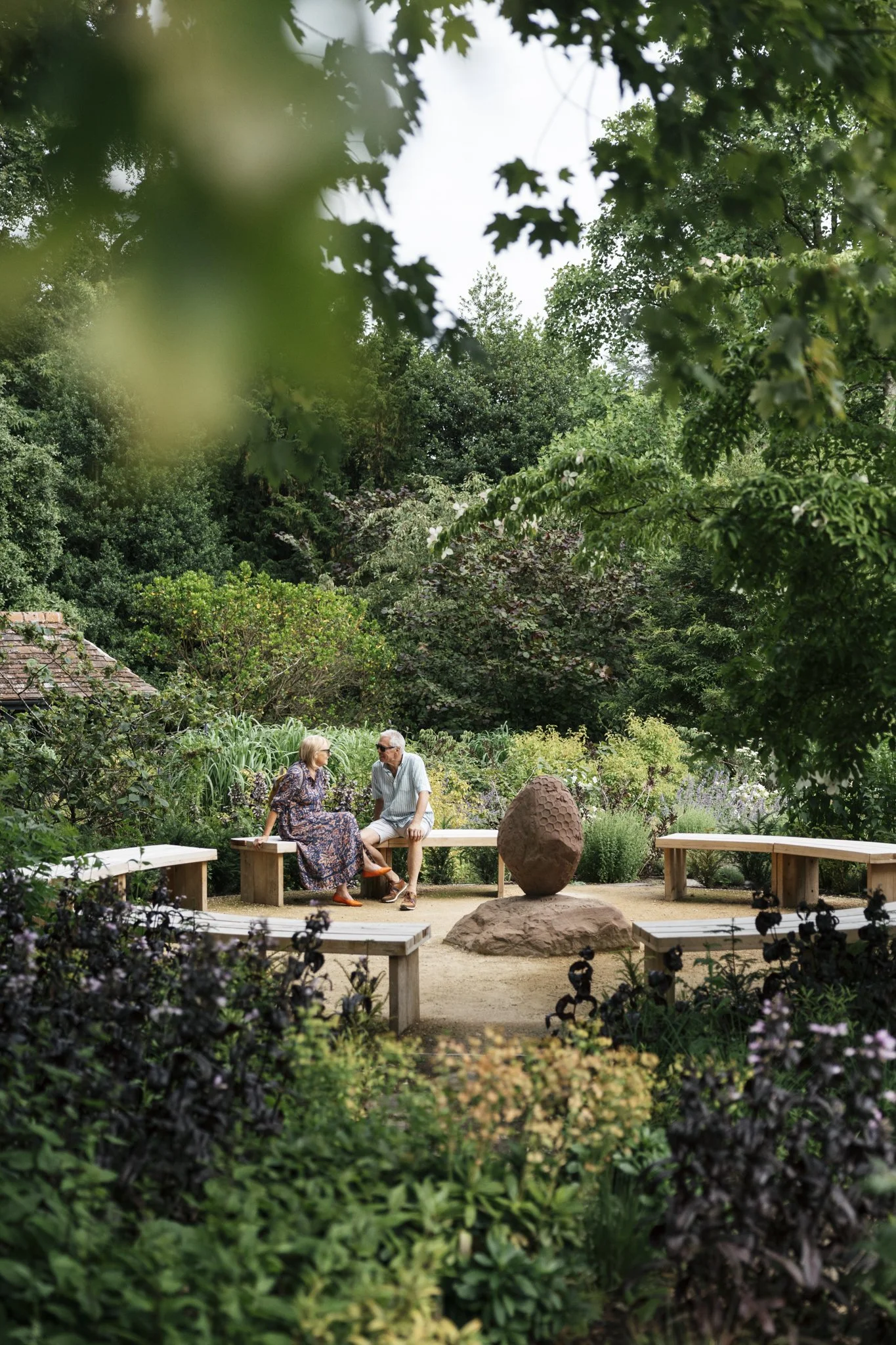 Two elderly people sitting and talking on wooden benches in a lush garden with abundant green plants and trees, centered around a large sculpture resembling a rock or egg.