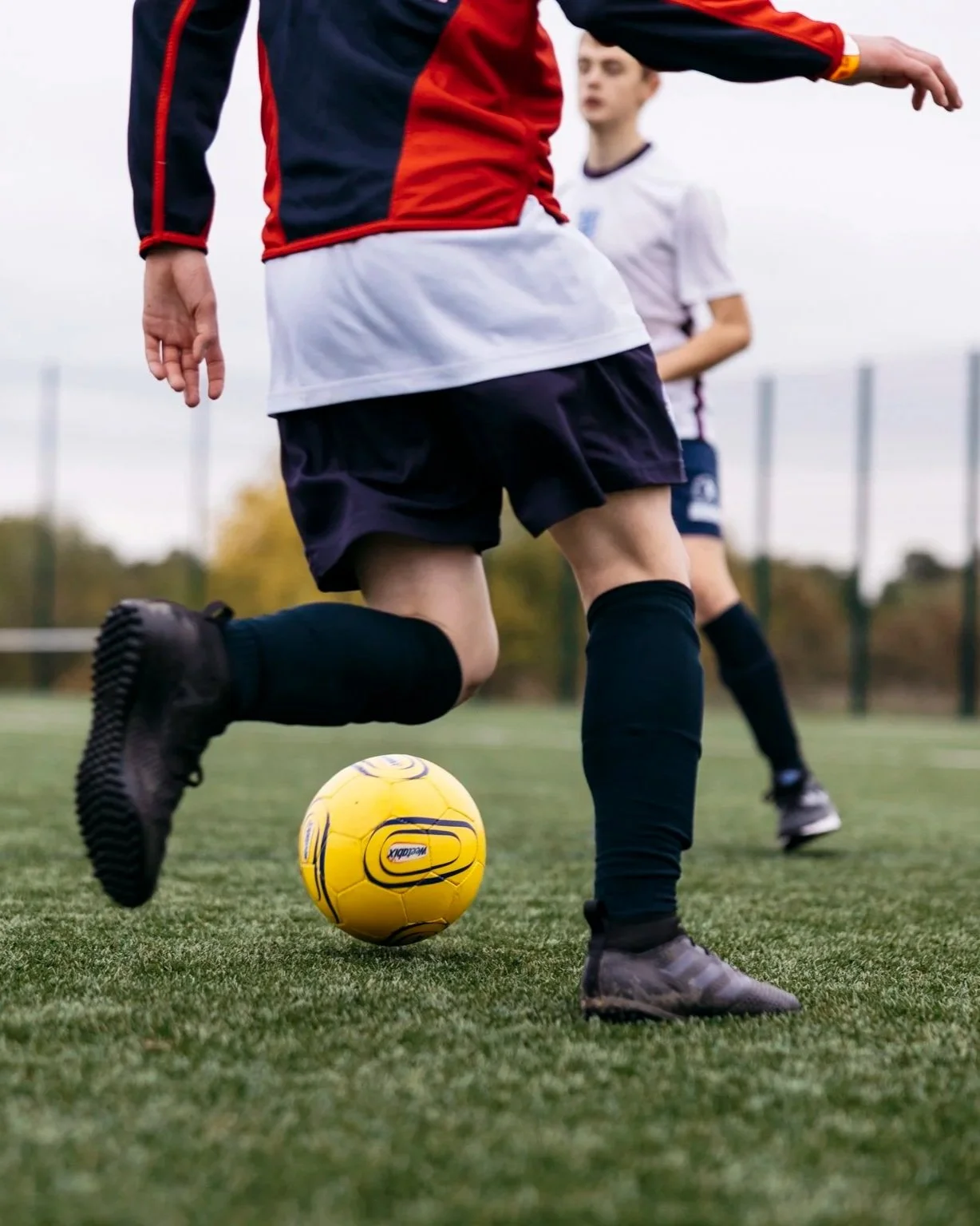 Soccer players in black uniforms with knee-high socks, one player is about to kick a yellow soccer ball on a grass field. Another player is in the background.