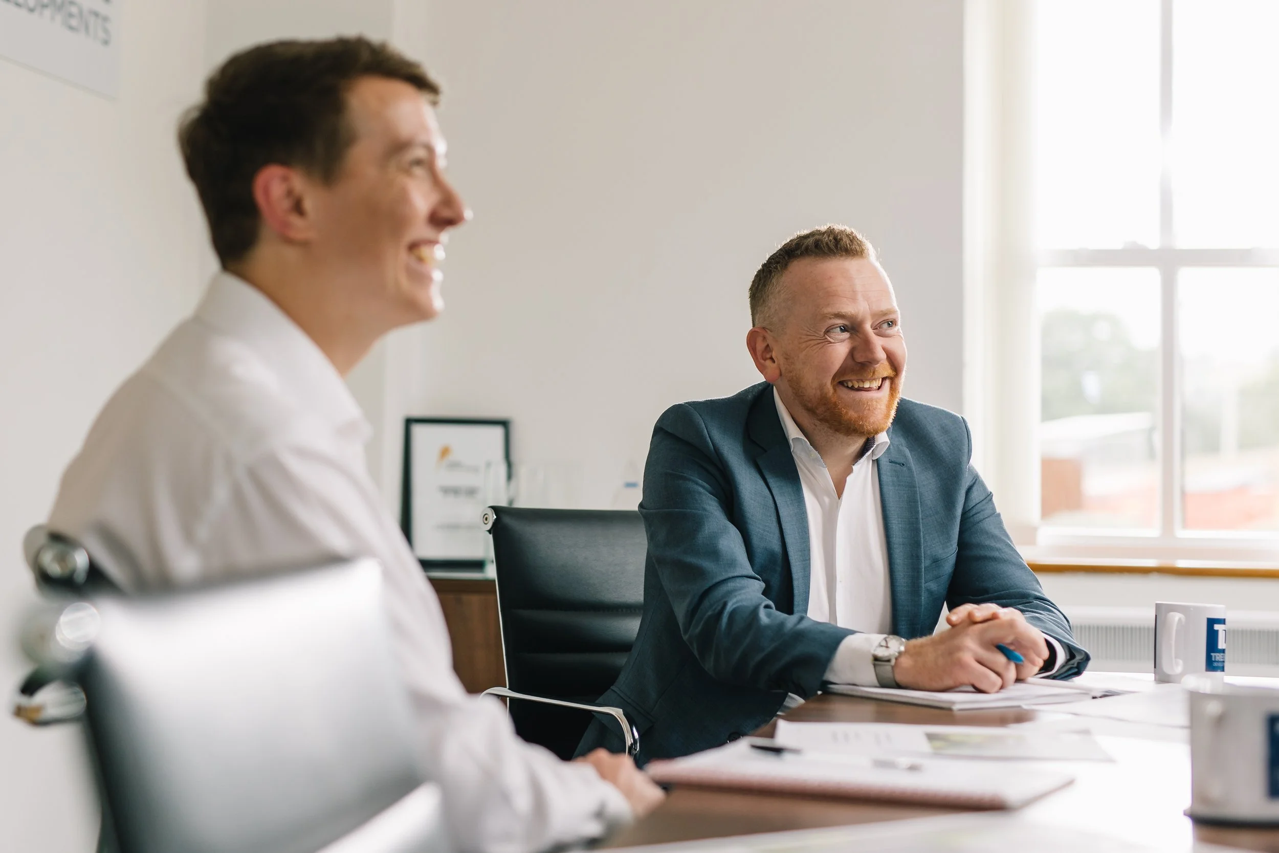 Two men sitting at a desk in a bright office, smiling and engaging in a conversation, with papers and mugs on the desk.