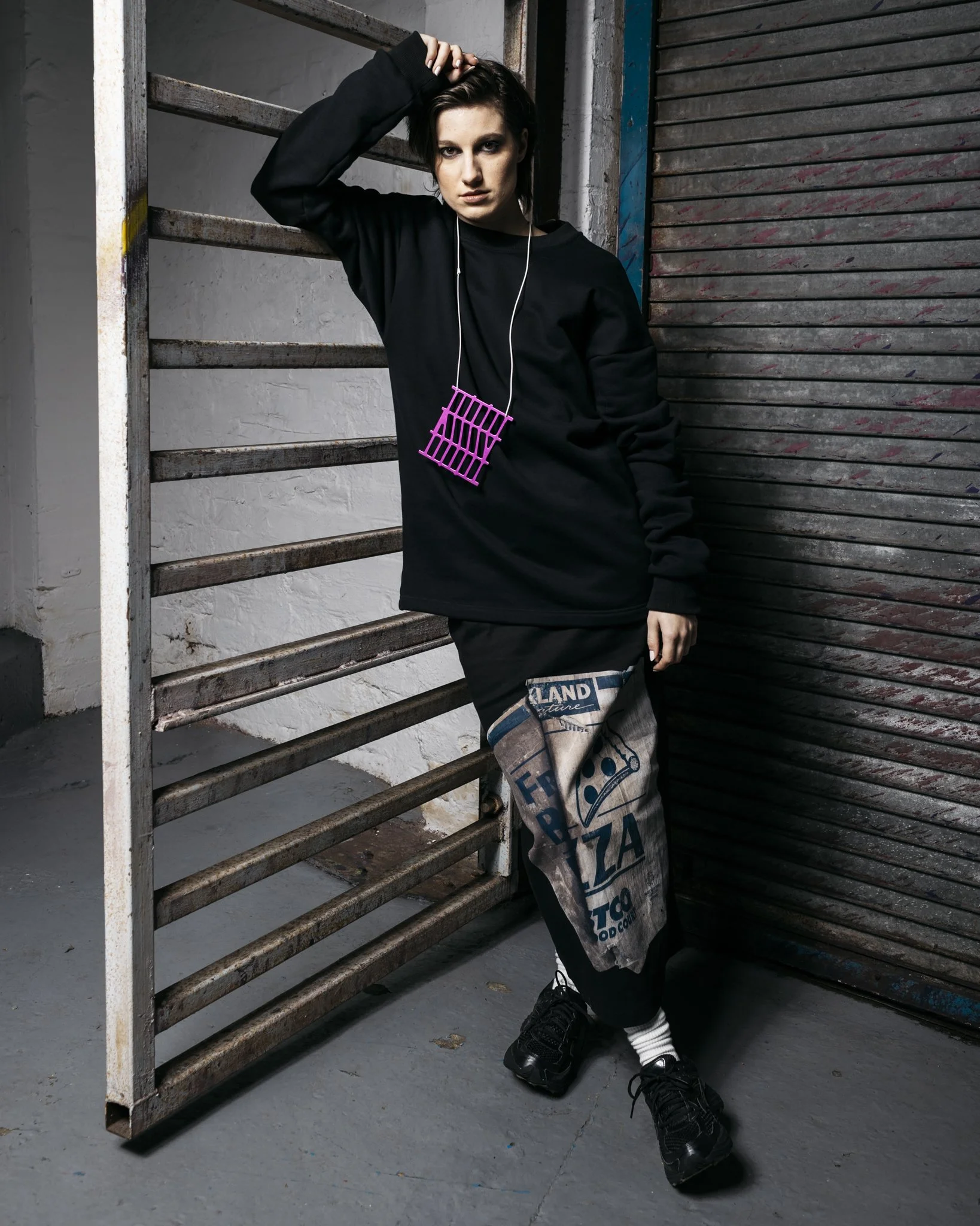 A woman in a black sweatshirt and black pants stands against a rustic metal gate and a textured wall in an industrial setting, wearing white earphones and holding a pink plastic card or object on a lanyard.