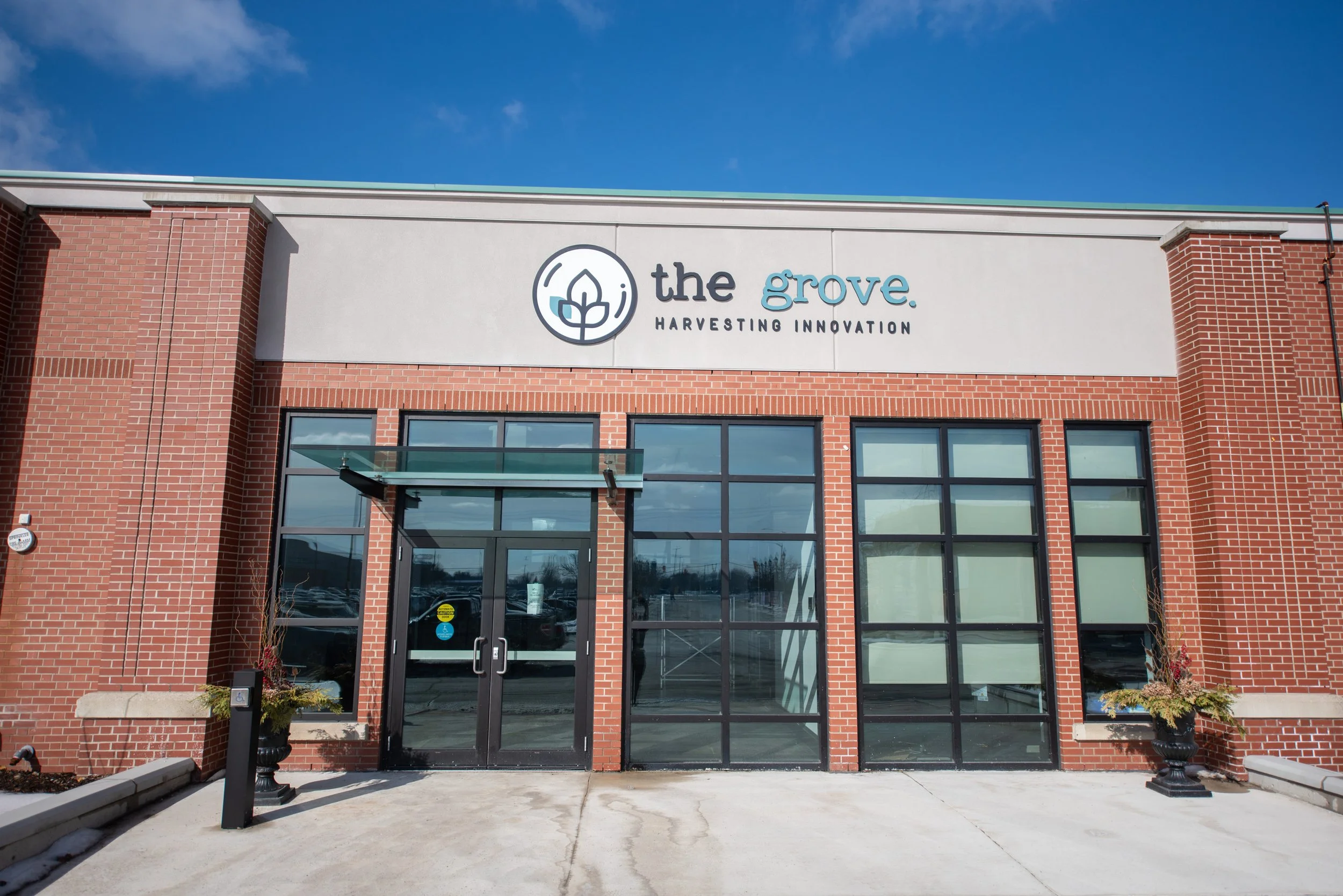The exterior of a brick building with a sign that reads 'the grove. HARVESTING INNOVATION,' with a logo of a leaf inside a circle. The entrance has glass doors and large windows, with small planters and a wheelchair accessible sign near the door.