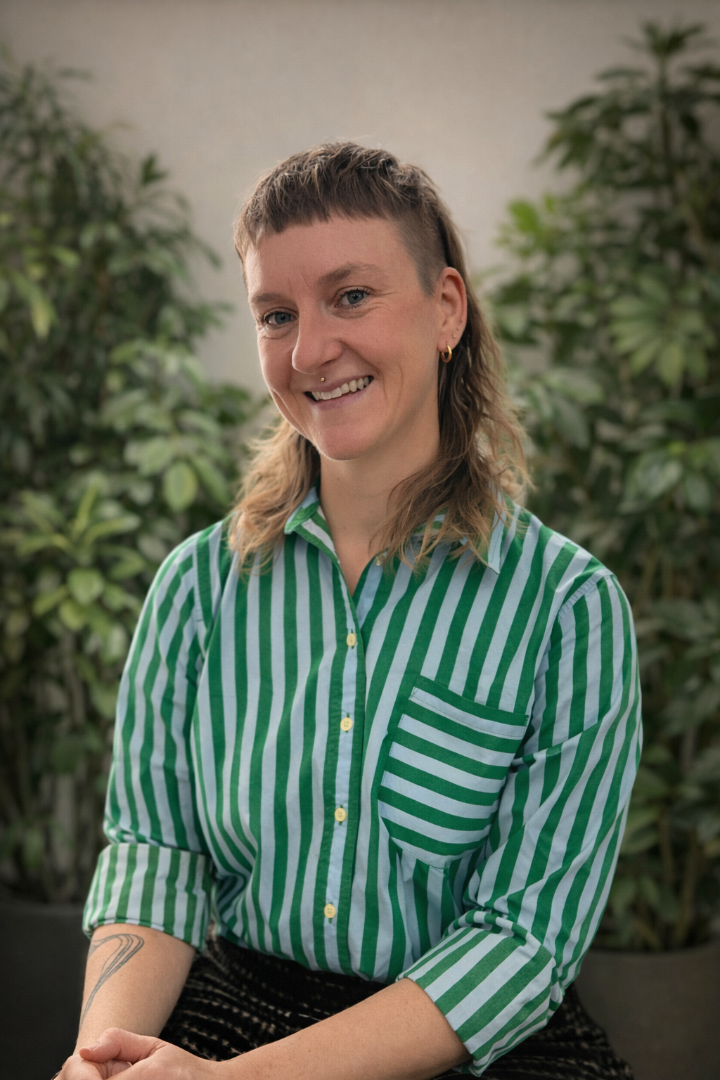 Image of Laura Foxx seated in a therapy office looking queer with a mullet and medusa piercing, wearing a green and blue striped shirt.