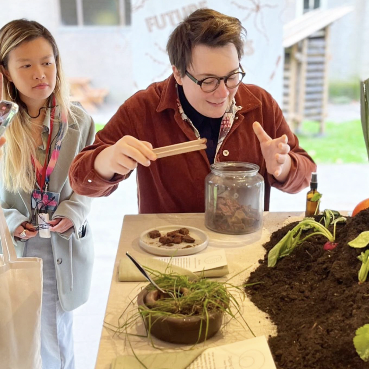 A person with glasses and brown hair is planting seeds or seedlings into soil on a table, with a jar and plates of small rocks nearby. A girl with long blonde hair and a gray jacket is observing. The background suggests an outdoor or semi-outdoor setting with a banner or sign.