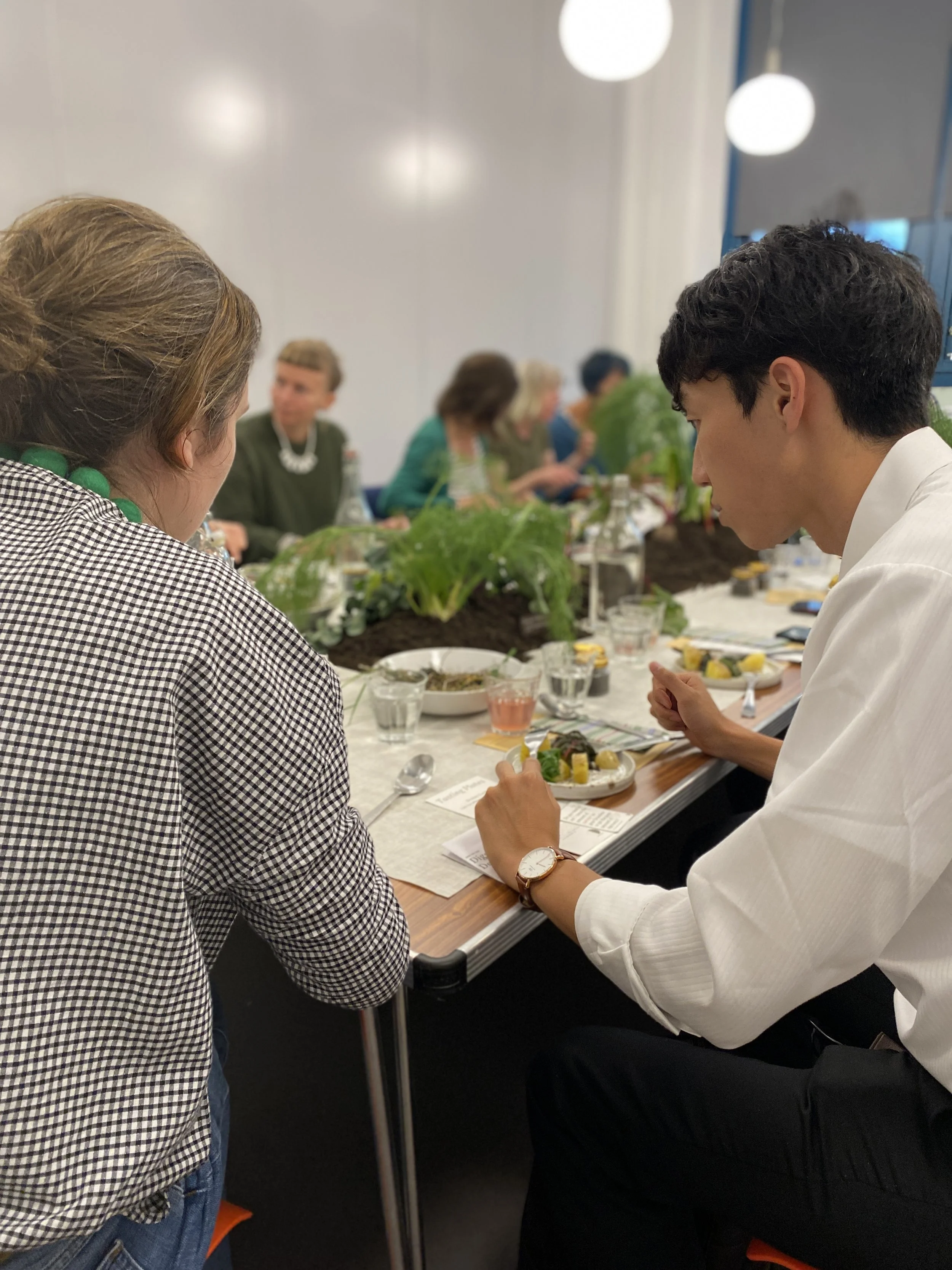 People gathered around a table with plates of food, glasses, and green plants, in a bright conference room, engaged in conversation.