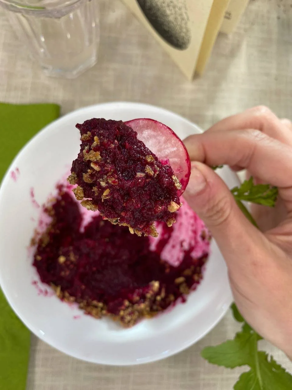 Hand holding a spoonful of berry cobbler with a slice of radish on top, over a white bowl filled with berry cobbler.