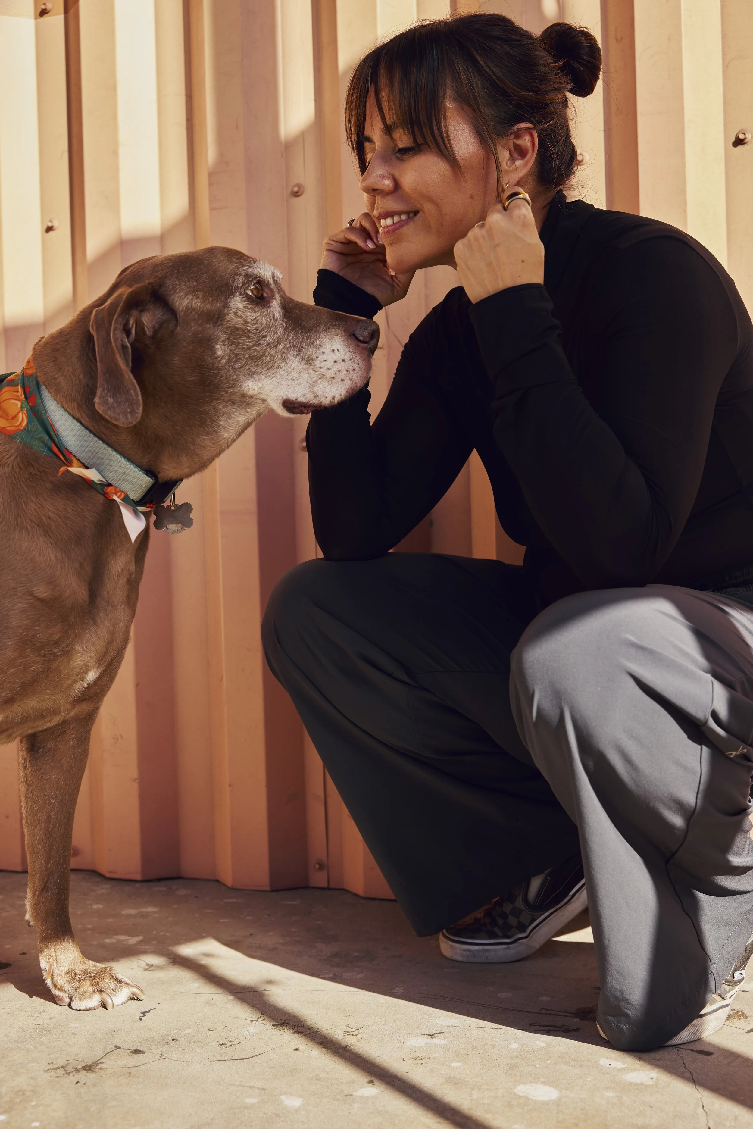 A woman in a black long-sleeve top and gray pants crouches down facing a brown dog with a colorful collar. The woman is smiling and touching her face near her ear while the dog looks at her. They are in front of a wooden wall with sunlight casting shadows.