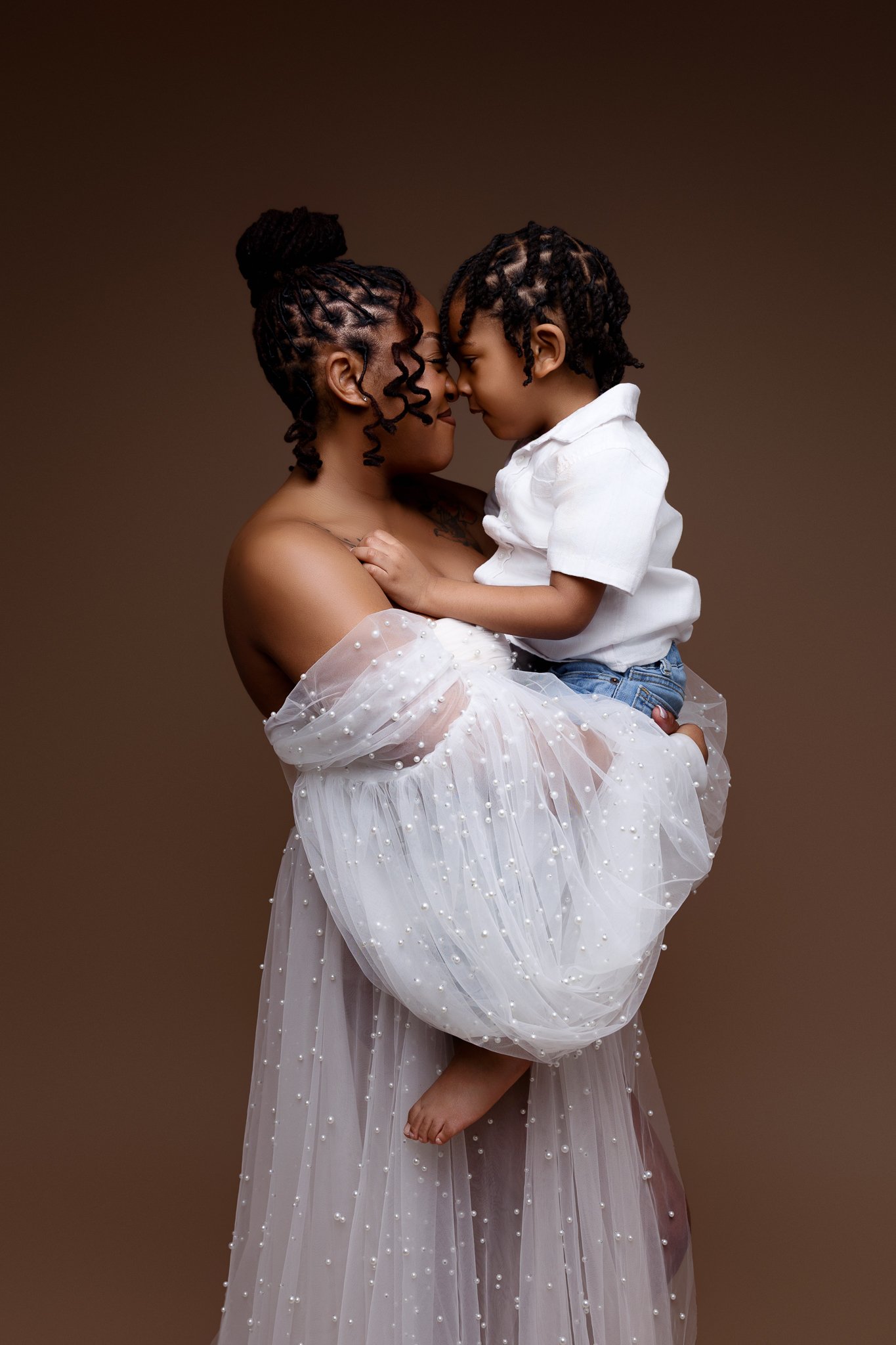 A woman with braided hair holding a young boy with braided hair. They are touching foreheads against a neutral brown background.