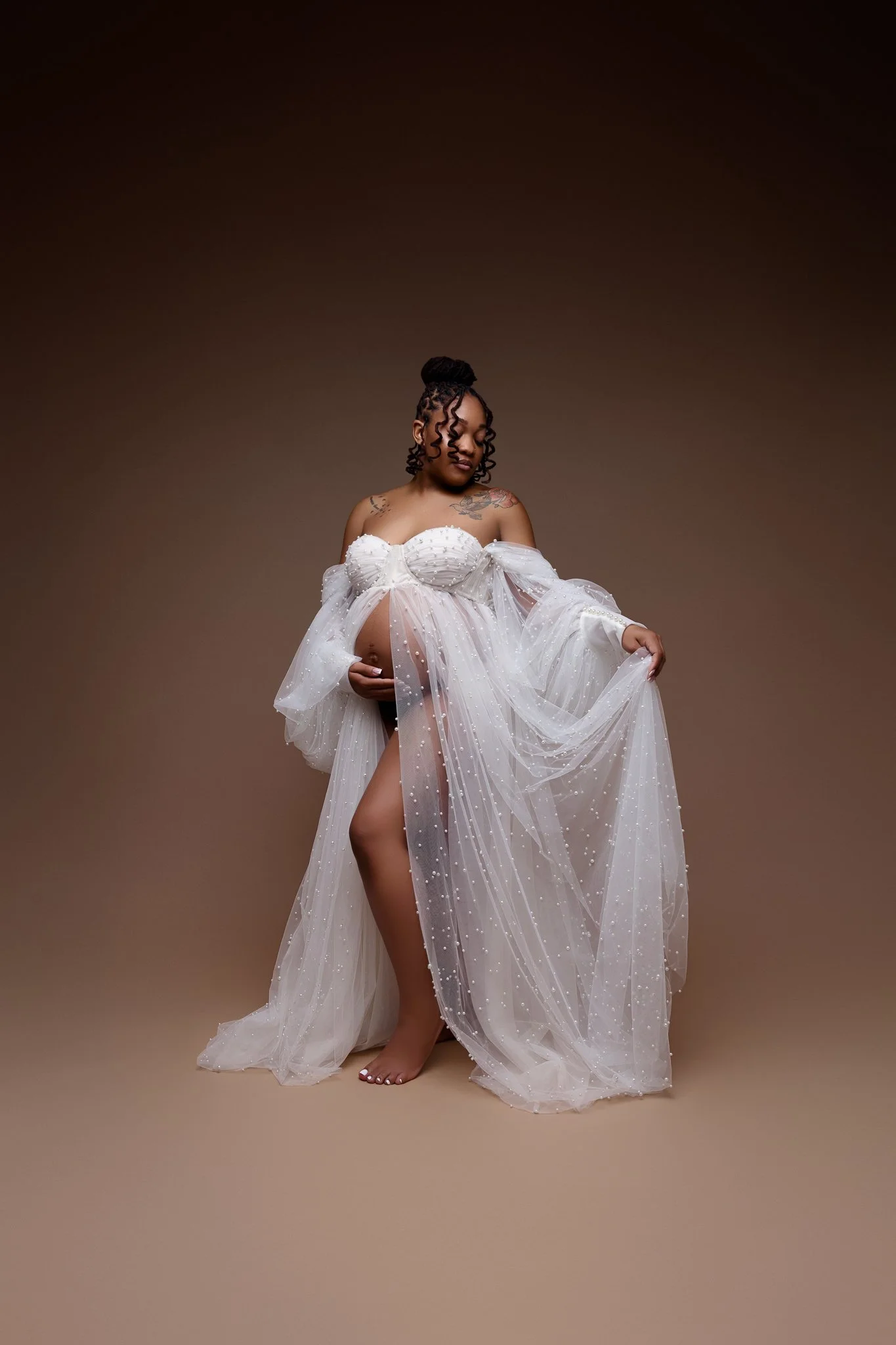 A pregnant woman in a sheer, white, embellished gown with off-the-shoulder sleeves, standing barefoot against a plain brown background. She has tattooed shoulders, curly hair styled in a bun, and is gently holding her baby bump.