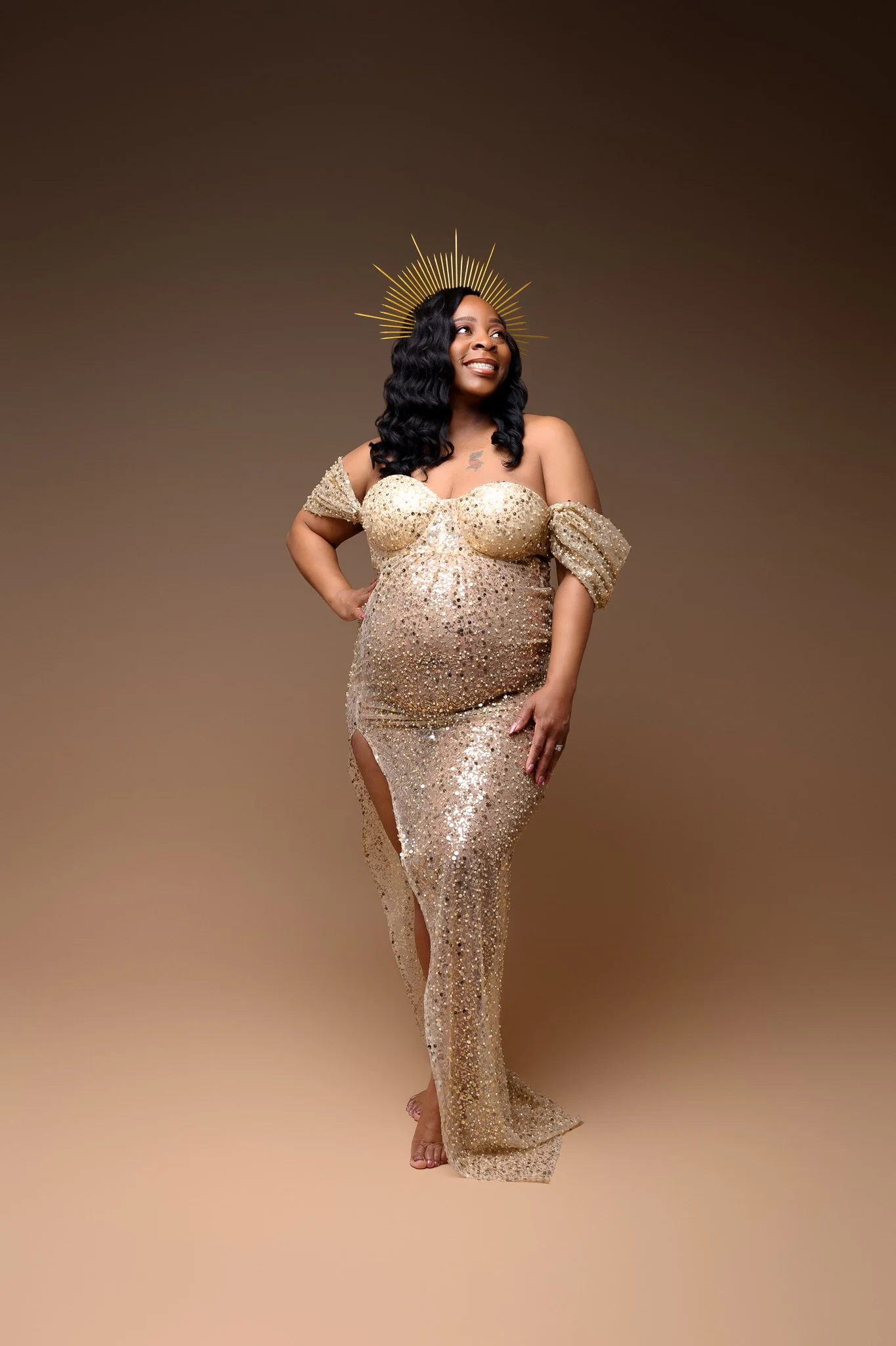 A woman wearing a strapless, gold, sequined gown with thigh-high slit, standing barefoot with a smile, and wearing a golden halo crown, against a plain brown background.