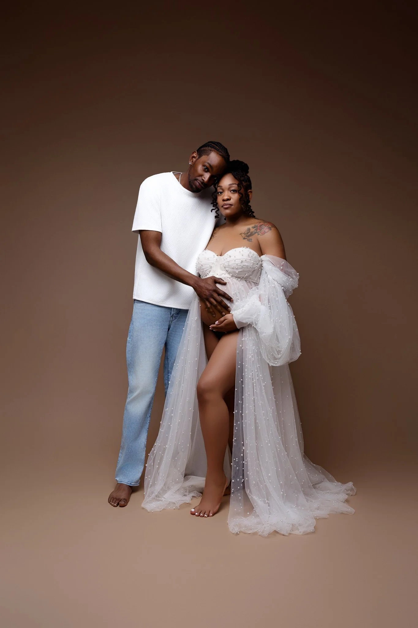 A pregnant woman in a sheer, white, star-embellished dress with off-shoulder puffy sleeves, standing barefoot on a beige background, with a man in a white t-shirt and light blue jeans placing his hands on her belly.