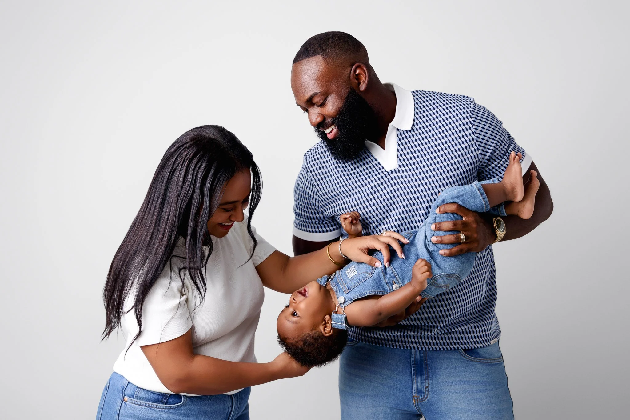 A mom and dad are playing with their toddler son in front of a white backdrop