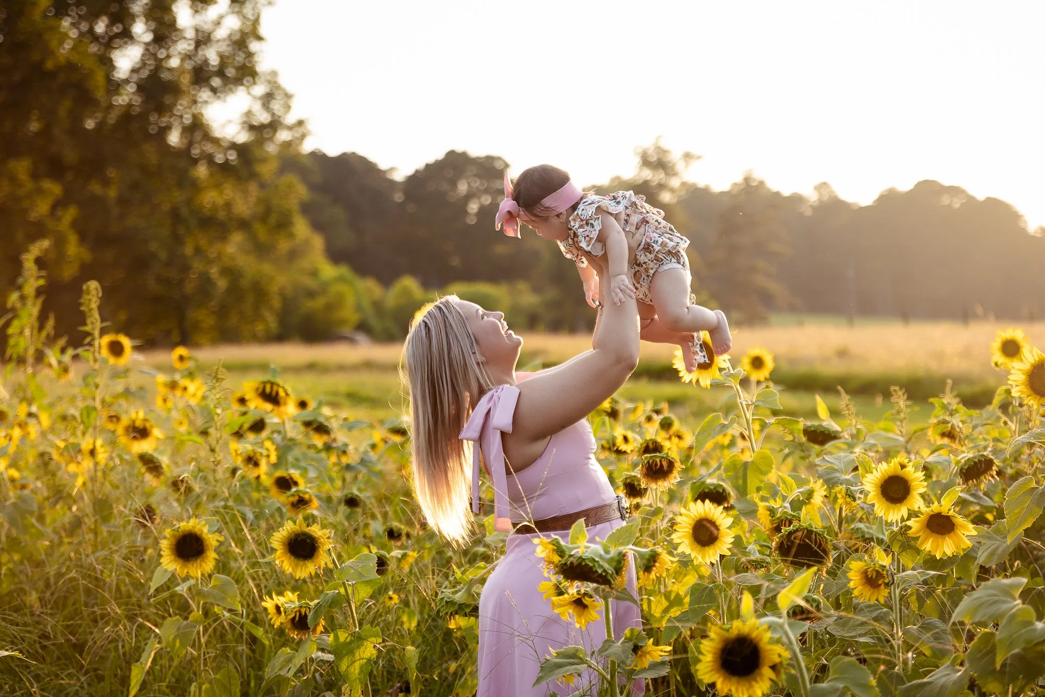 A woman lifting a young girl in a sunflower field at sunset in Atlanta, Ga, smiling and enjoying their time together.