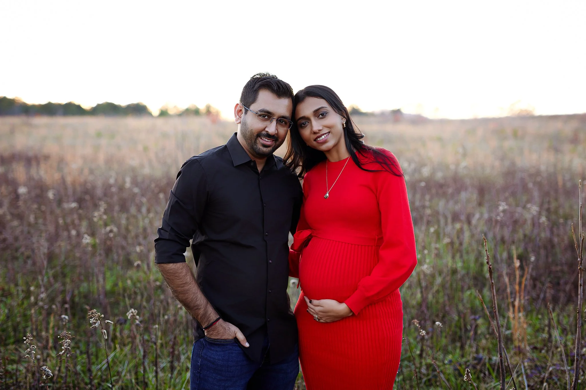 A young couple stands close together in a field during sunset in Atlanta, GA. The man is wearing glasses and a black shirt, and the woman is pregnant, wearing a red dress. Their heads are touching, and they are smiling gently at the camera.