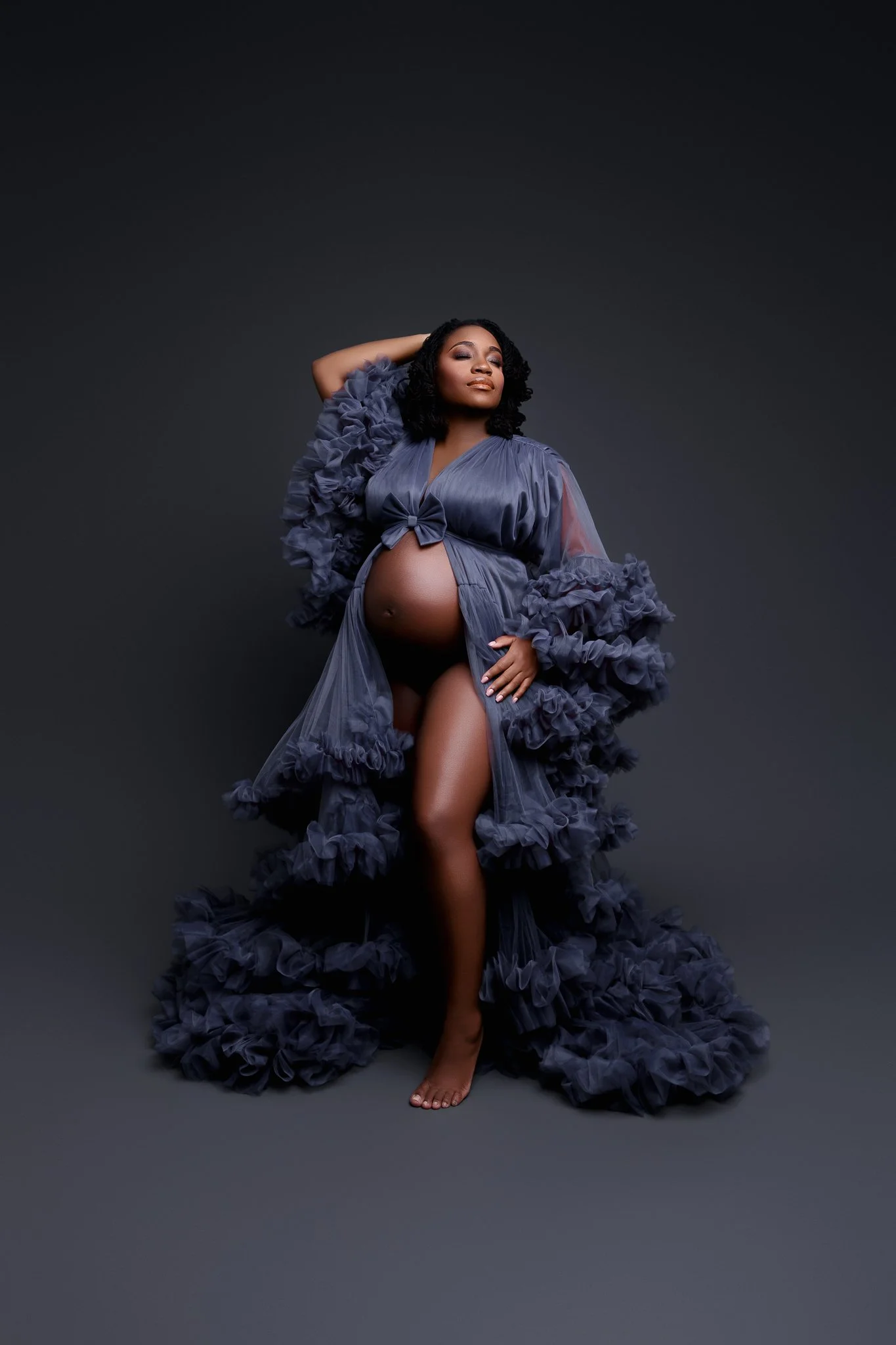 Pregnant woman with dark curly hair posing in a flowing, ruffled gray gown against a dark gray background.