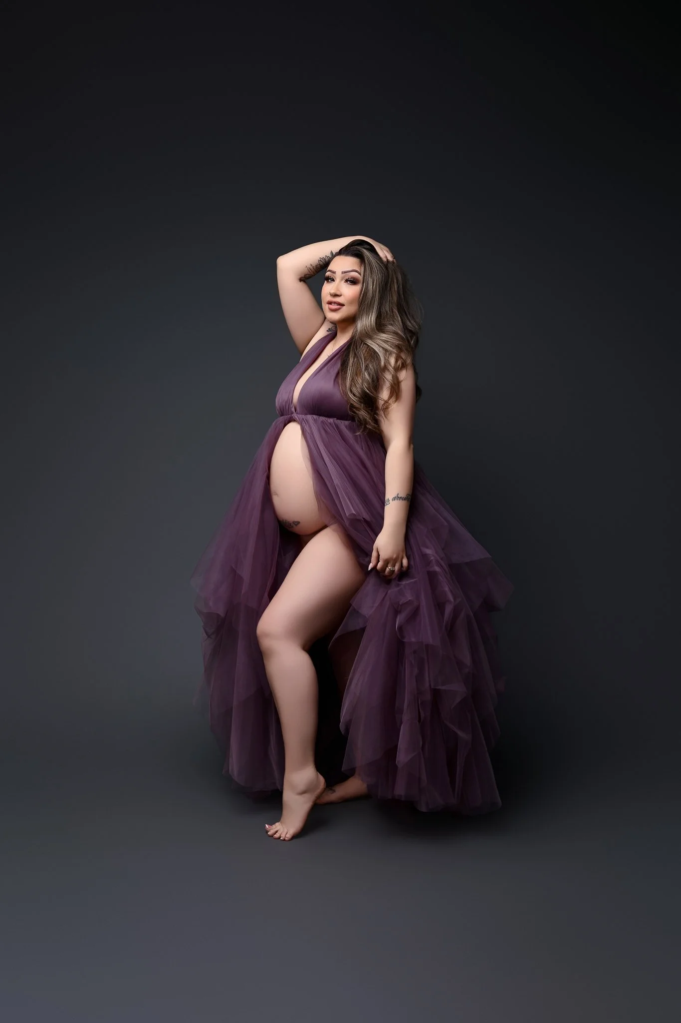 A pregnant woman is posed in a purple dress while ruffling her hair.