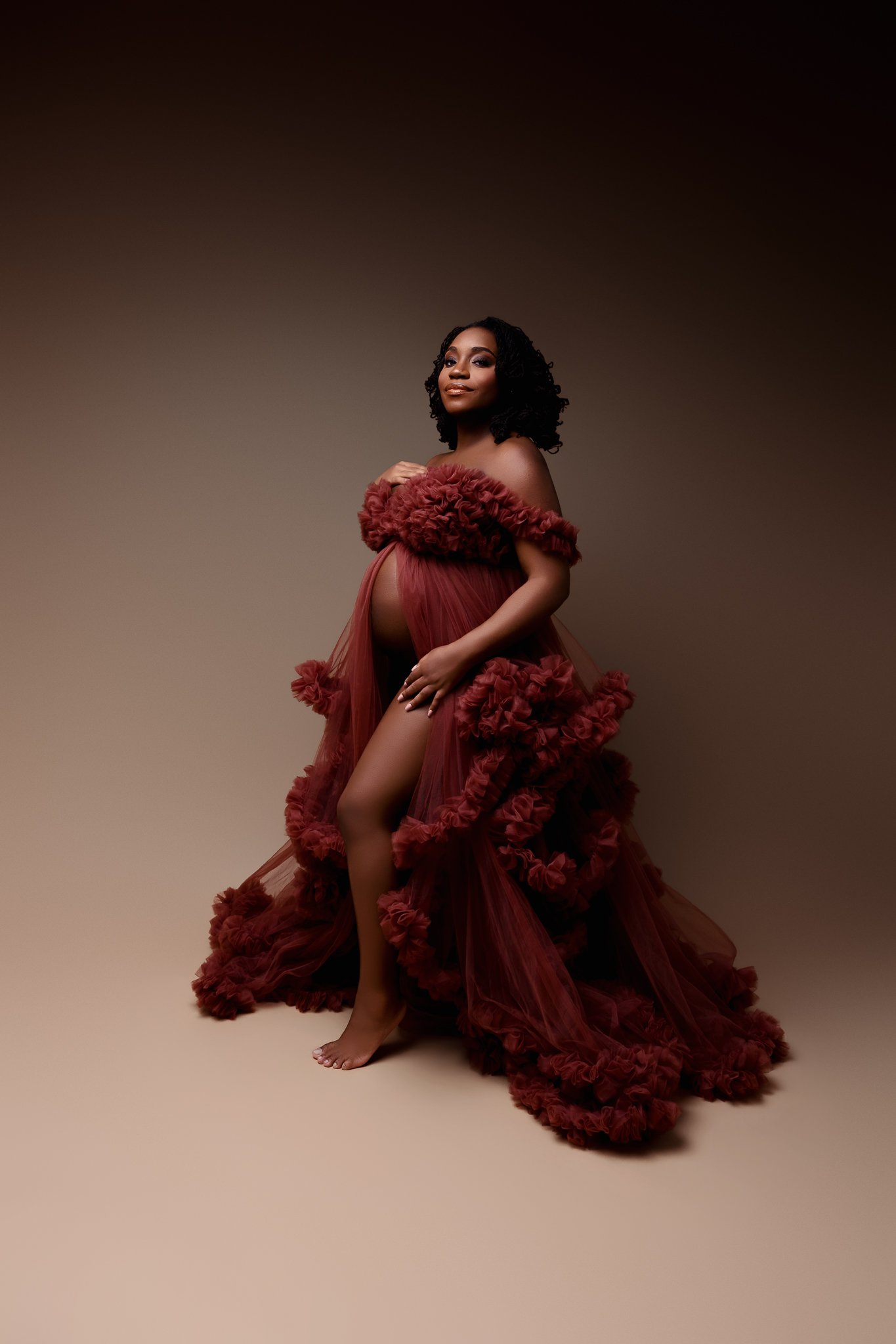 Pregnant woman wearing a flowing, ruffled, red dress with off-the-shoulder design, standing barefoot against a neutral gradient background.
