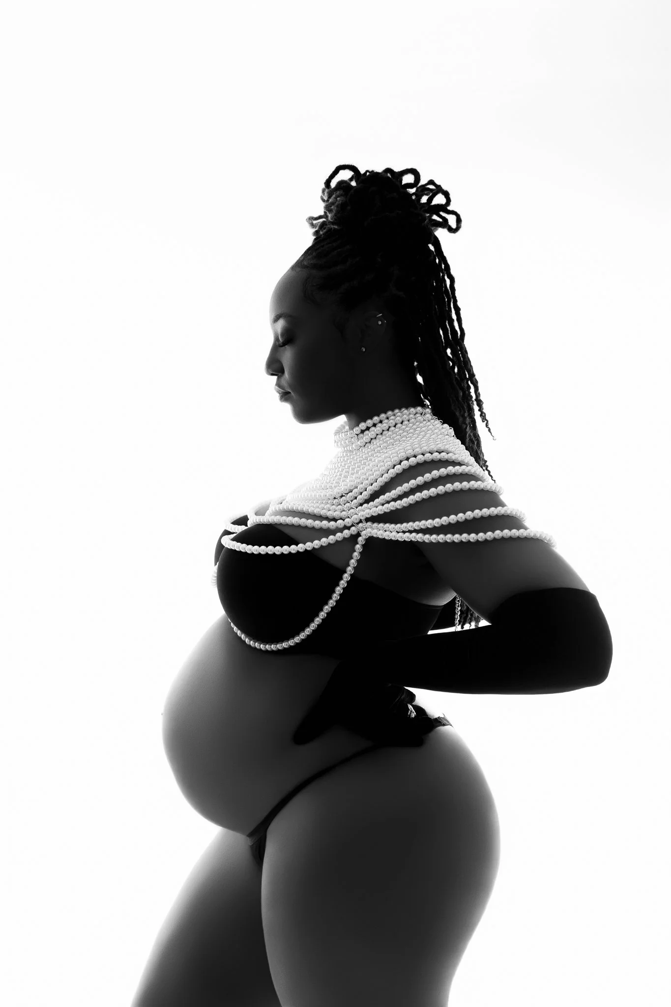 Profile of pregnant woman with dreadlocks, adorned with multiple strands of pearls, wearing black lingerie and long gloves, against a white background.