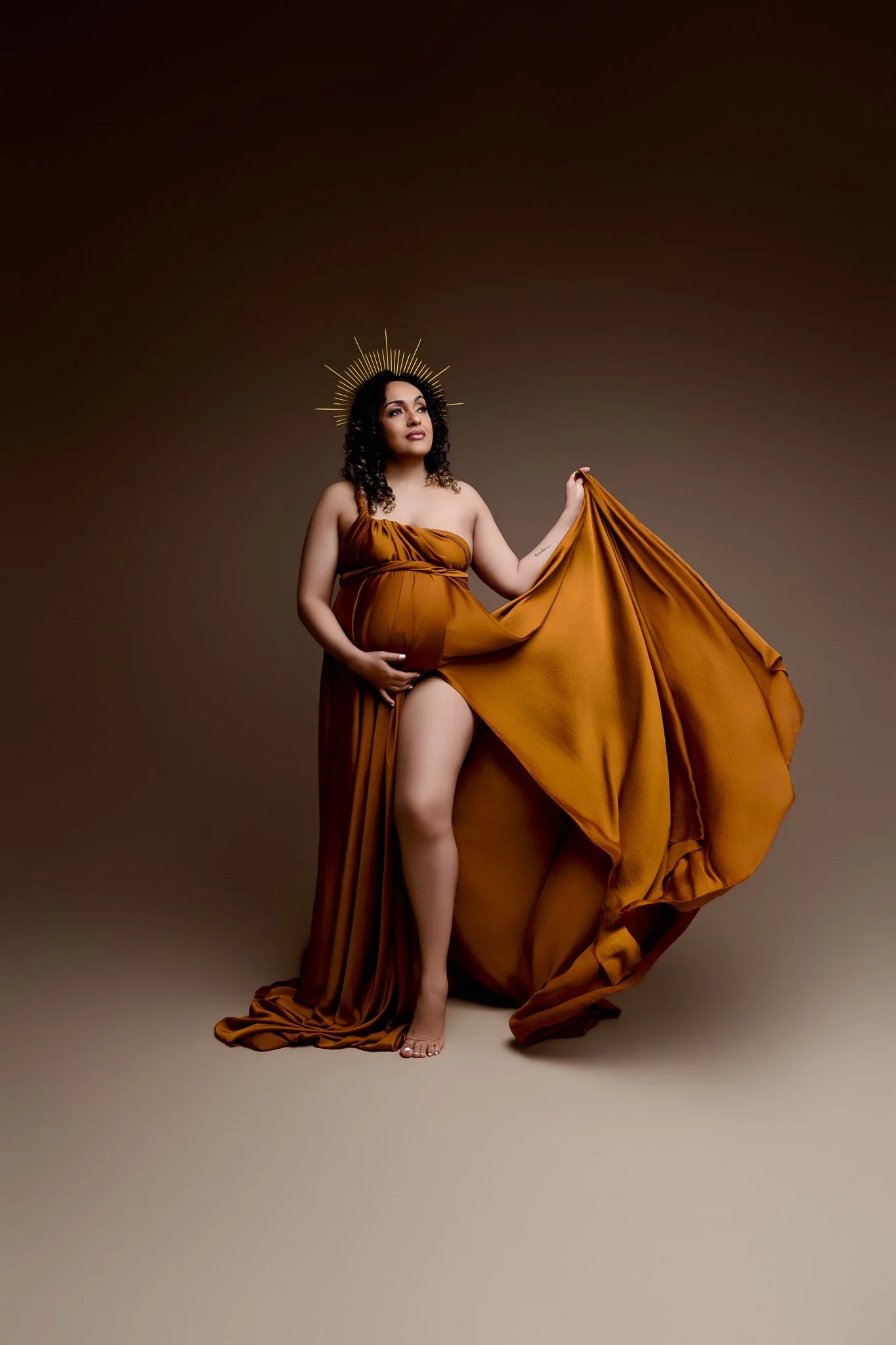 A pregnant woman wearing a flowing, long, brown dress, holding her belly with one hand and lifting part of her dress with the other, standing against a plain, gradient background. She has curly hair and is adorned with a gold halo-like headpiece.