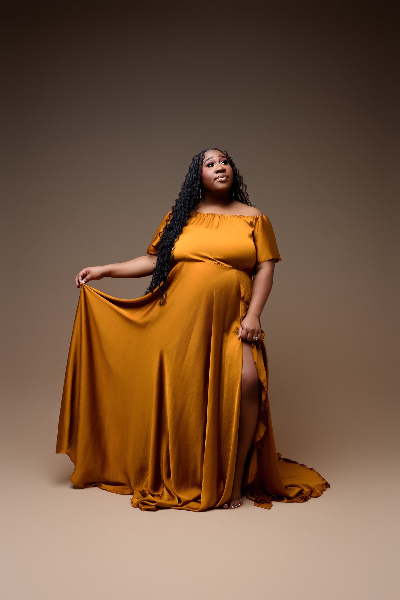 A woman with long, curly black hair, wearing a golden-brown satin off-the-shoulder gown, holding the dress to reveal one leg and standing against a plain gradient background.