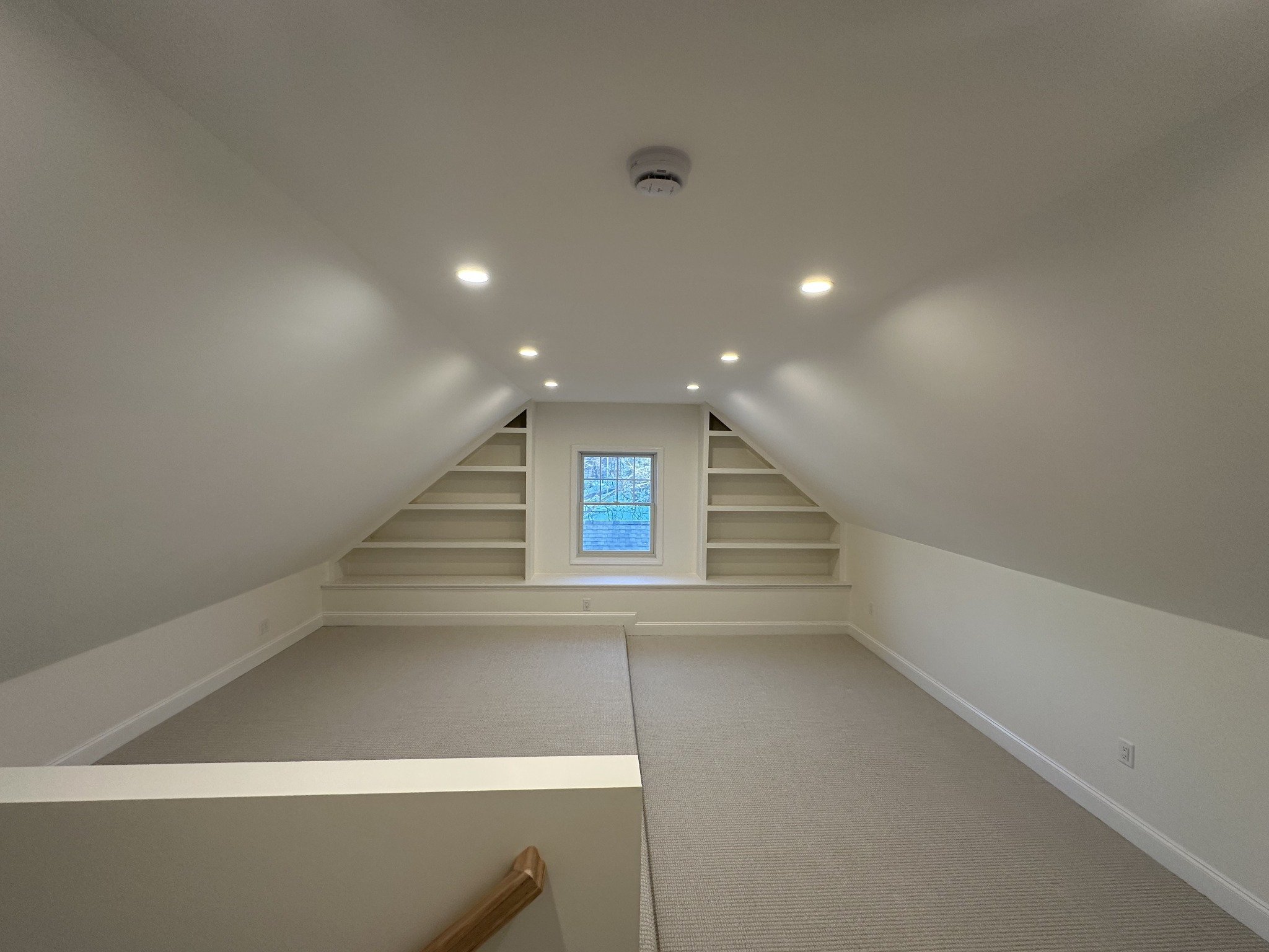 Finished attic with carpet, recessed ceiling lights and built-in shelving around the end window.