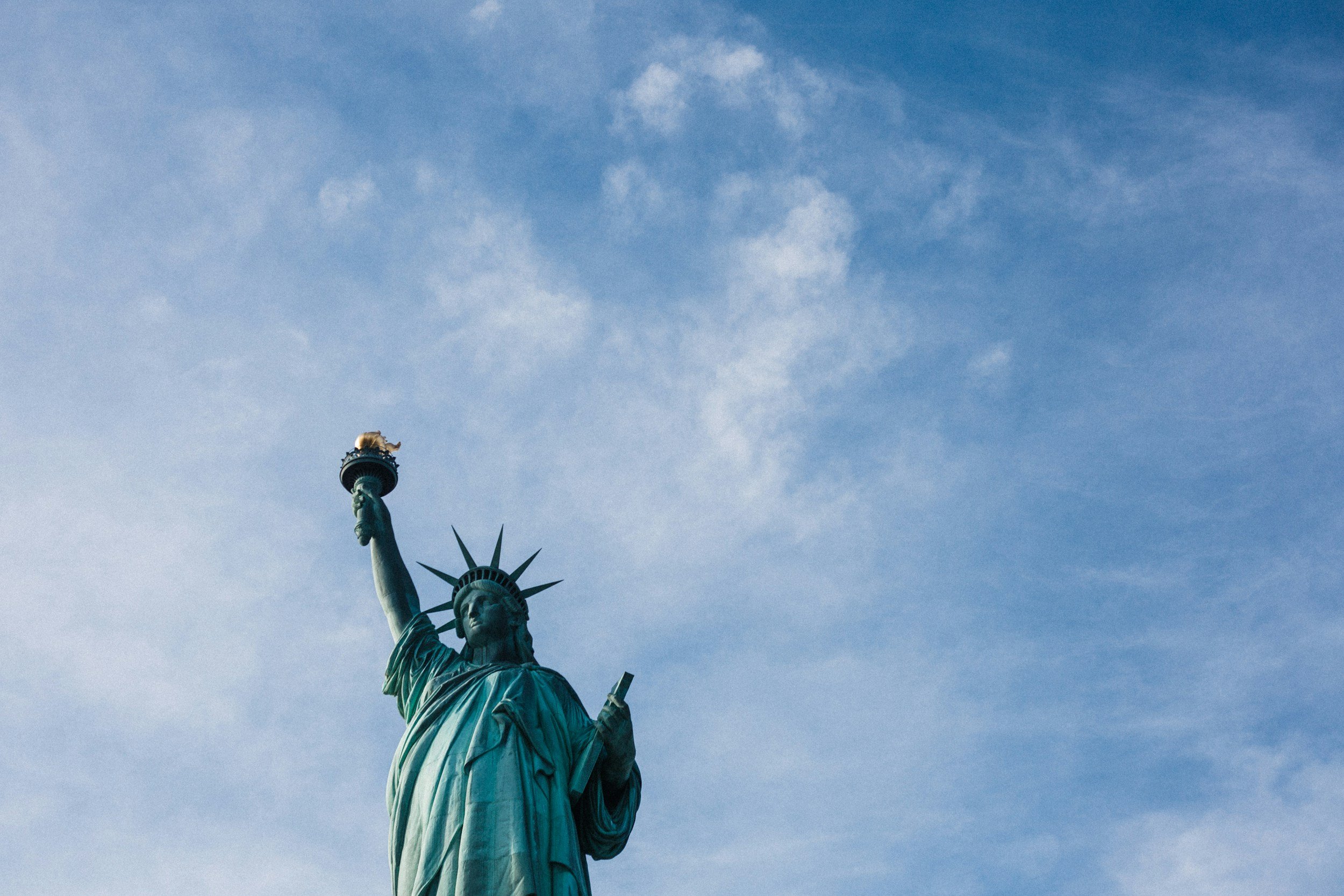 Top part of the Statue of Liberty held high against a blue sky with some scattered clouds.