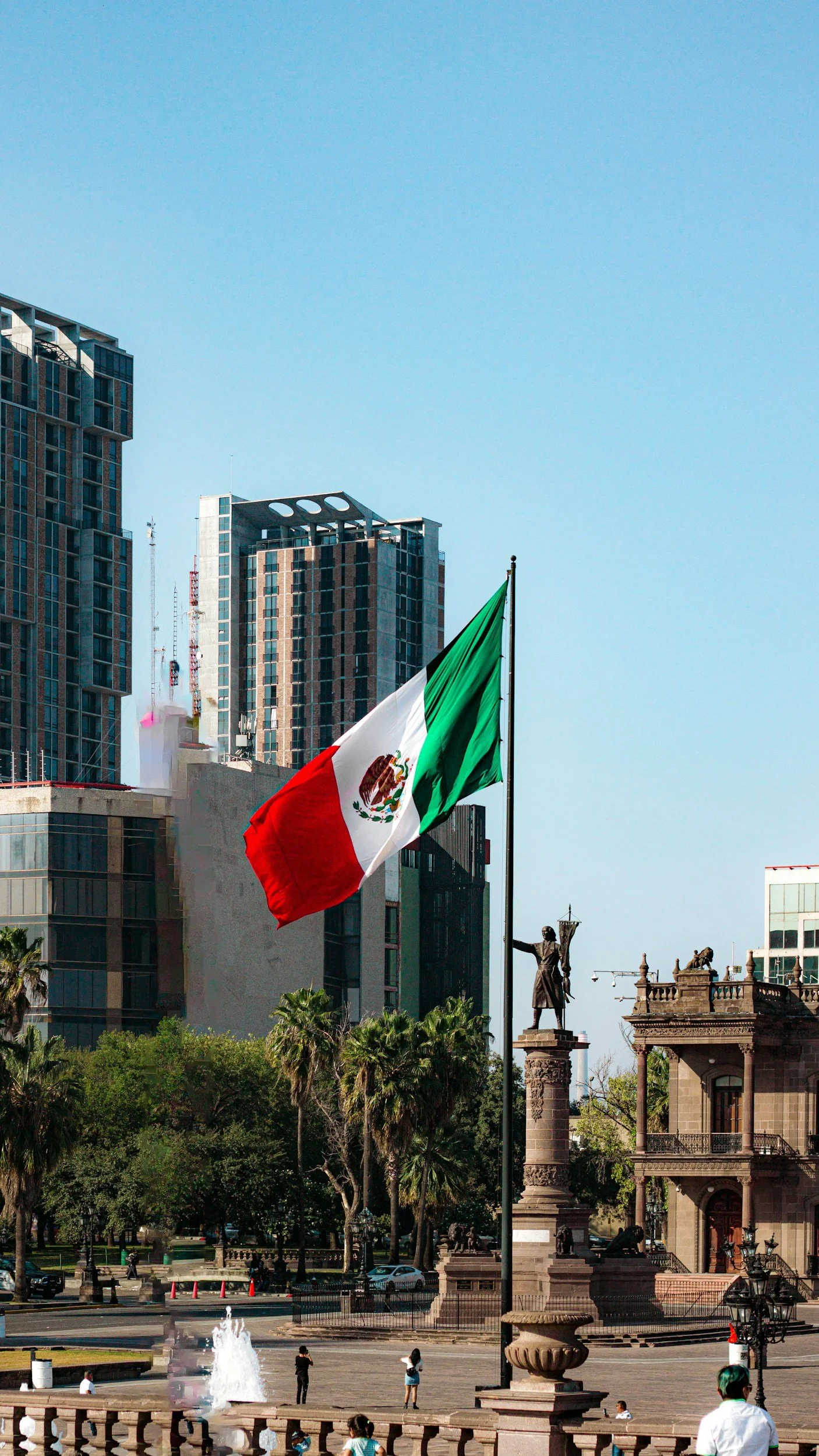 Mexican flag flying in a city square with modern skyscrapers and historic statues in the background.