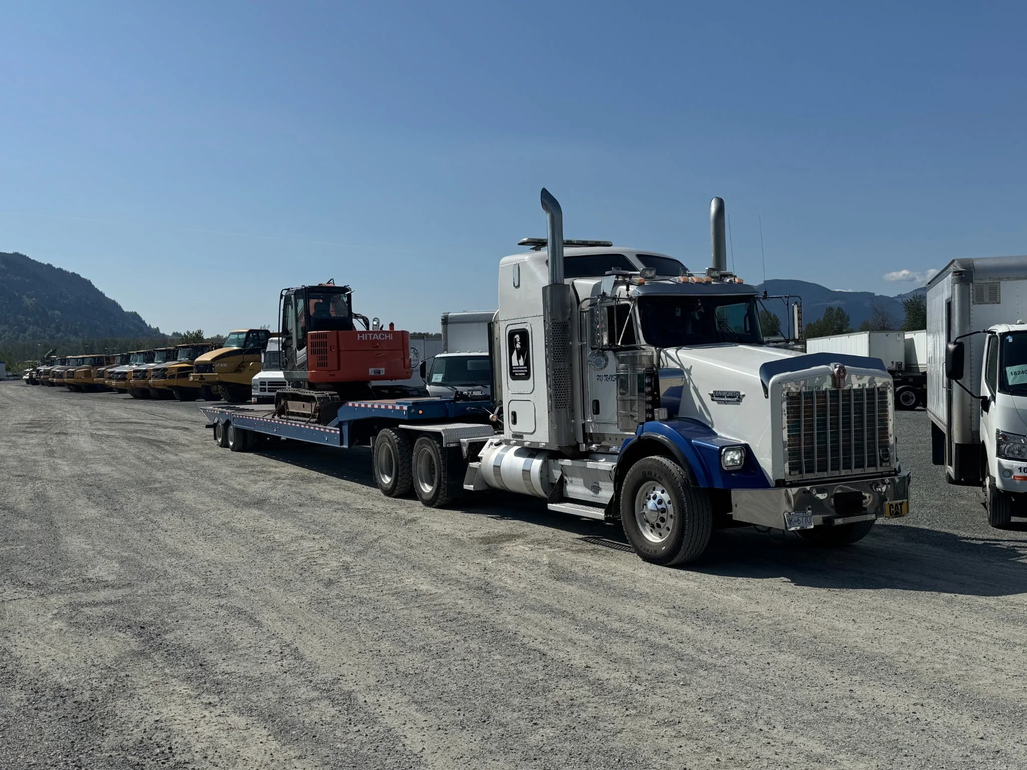 A white and blue semi-truck transporting a small orange excavator on a flatbed trailer, with several other trucks and construction vehicles parked in the background on a gravel lot, mountains and a clear sky in the distance.