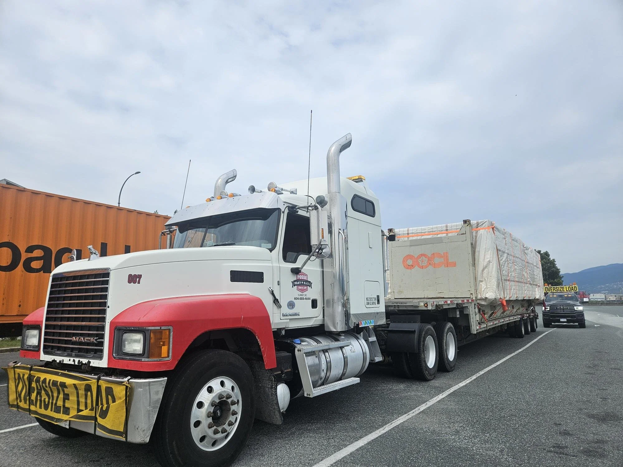 A white Mack semi-truck with red accents on the front, parked on the side of the road. The truck has a yellow sign that says "OVERSIZE LOAD" hanging from the front bumper, and a large cargo enclosed in clear wrapping on the flatbed trailer. There is a large orange container in the background and a dark-colored vehicle with a yellow "OVERSIZE LOAD" sign behind the truck.
