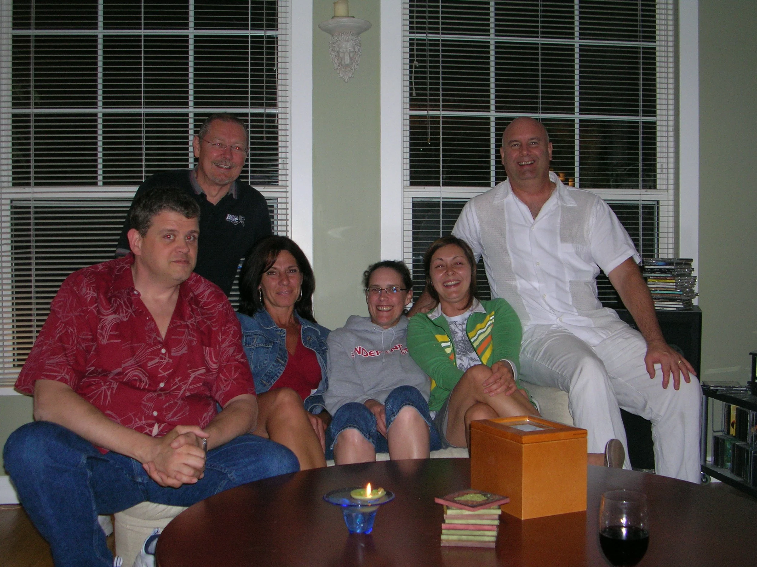 Group of eight people gathered around a table in a living room, smiling at the camera. There are two men standing and six people sitting, with a lit candle, a wooden box, and a stack of coasters or books on the table.