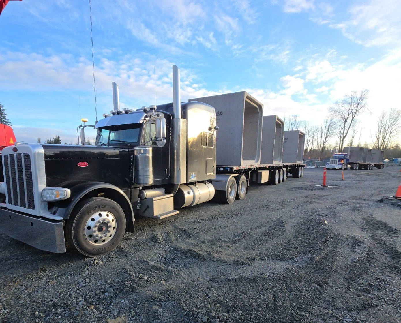 Black semi-truck with a flatbed trailer carrying large concrete blocks parked on gravel lot near trees and orange traffic cones, under partly cloudy blue sky with sunset glow.