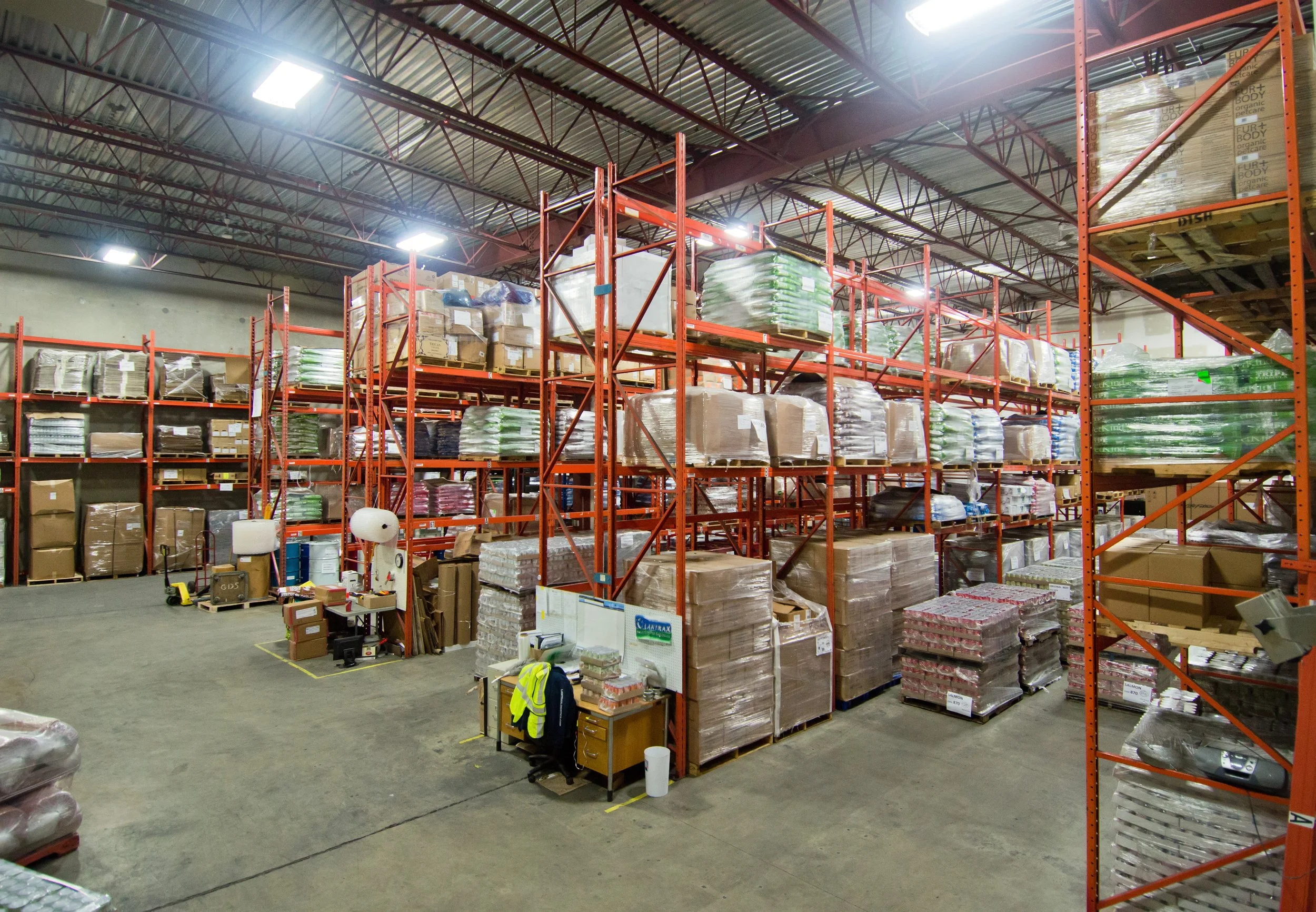 Interior view of a warehouse with tall orange metal shelving units filled with boxes and packages, overhead lighting, concrete floor, and a high ceiling with metal beams.