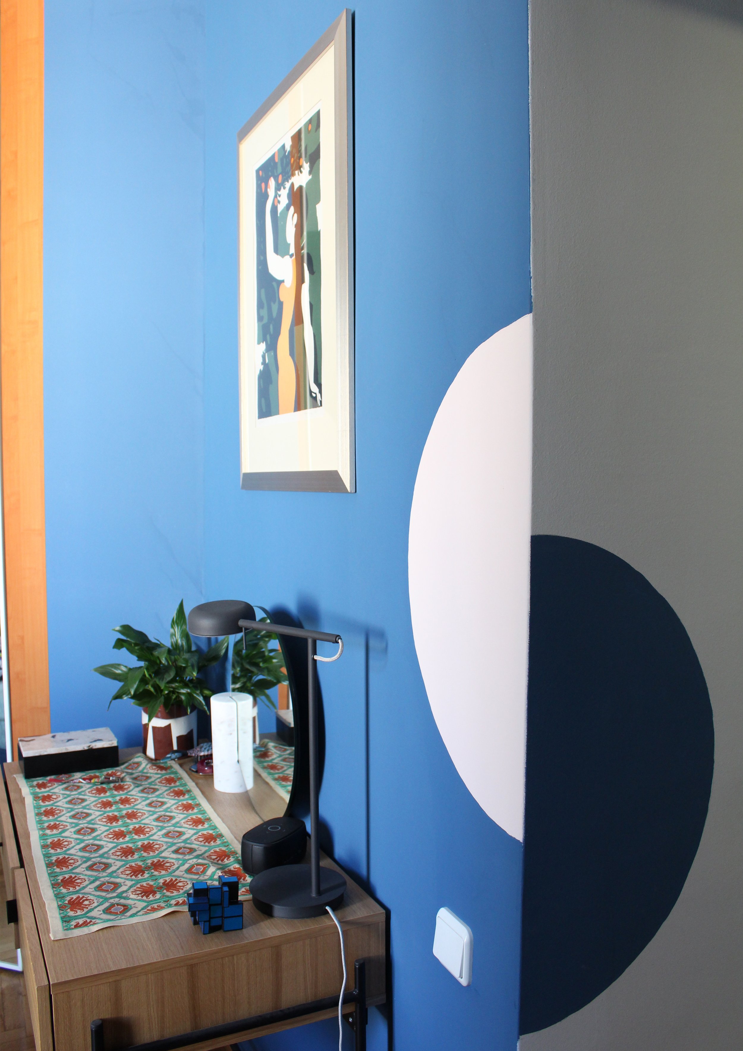 A room corner with a blue wall featuring a large black and white circular design, a framed abstract art piece, a wooden desk with a table lamp, a rubik's cube, some potted plants, and various small items including tissues and a box.