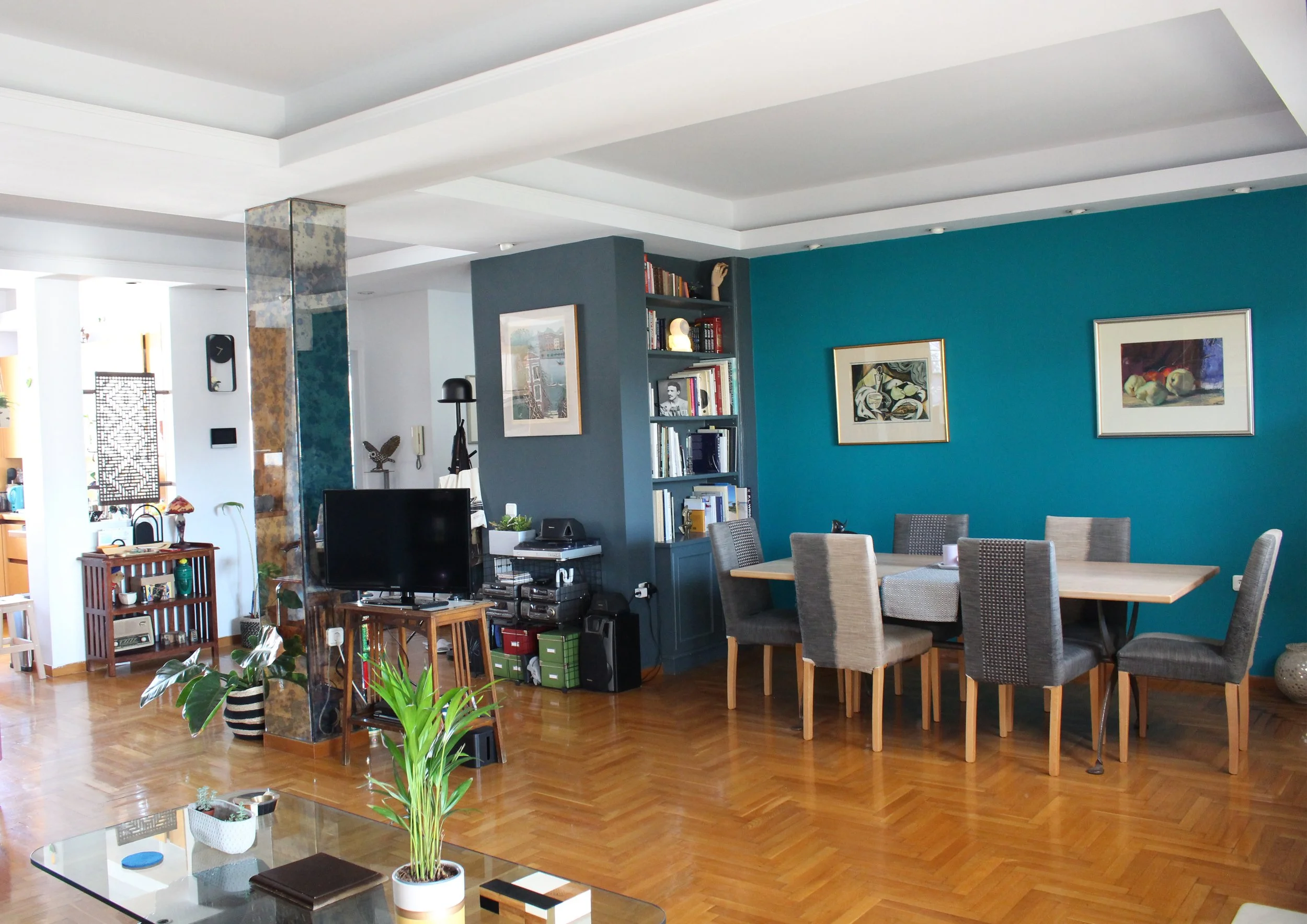 Interior view of a living and dining area with a teal accent wall, framed artwork, a dining table with six chairs, a television, bookshelves, and various decorative items, with wooden flooring.