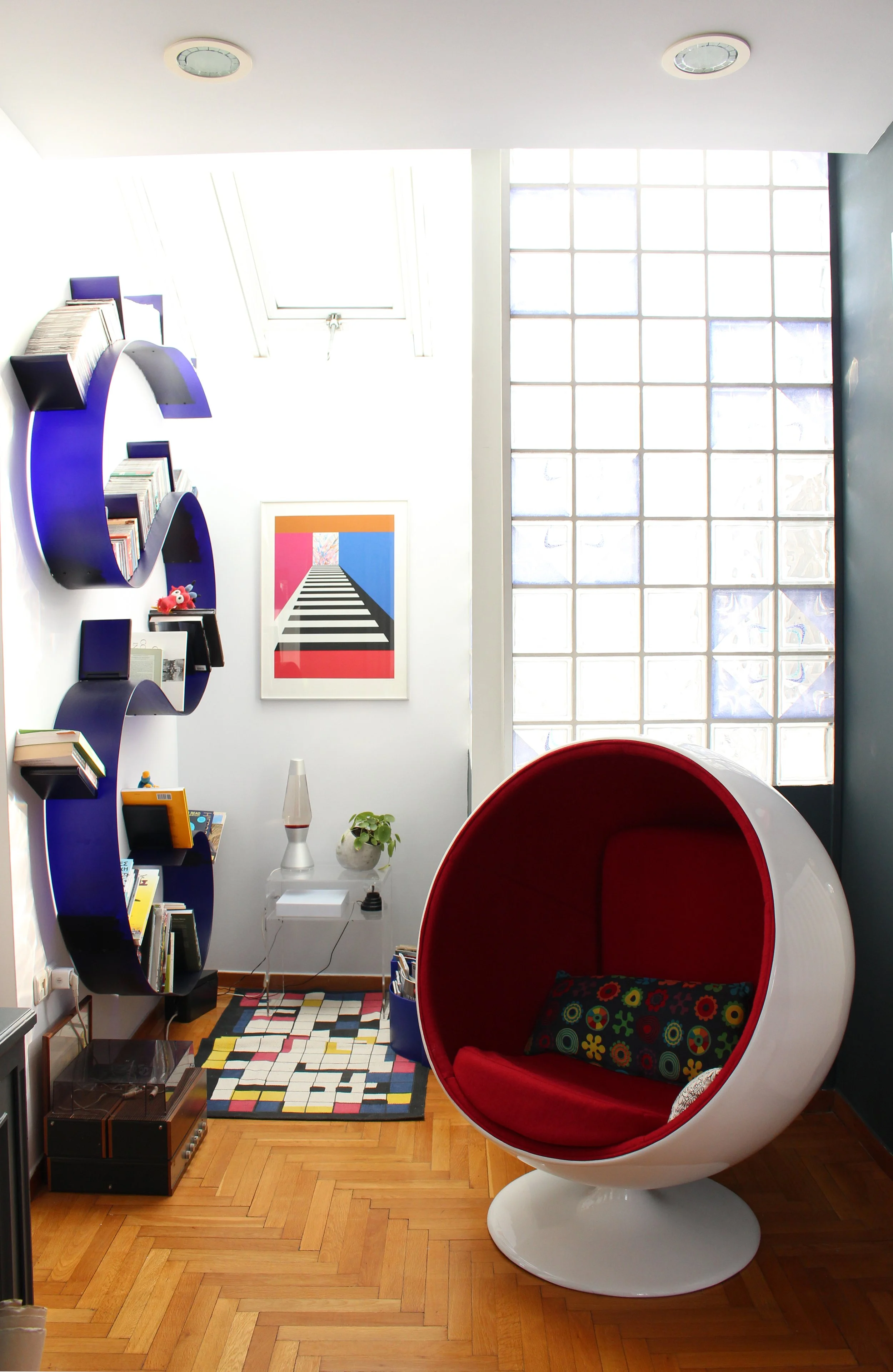 Modern interior room with white and black walls, wooden floor, red and white spherical lounge chair, colorful patterned cushion, geometric multicolor rug, bookshelf with books, framed artwork, small table with plants, lava lamp, and a glass block wal