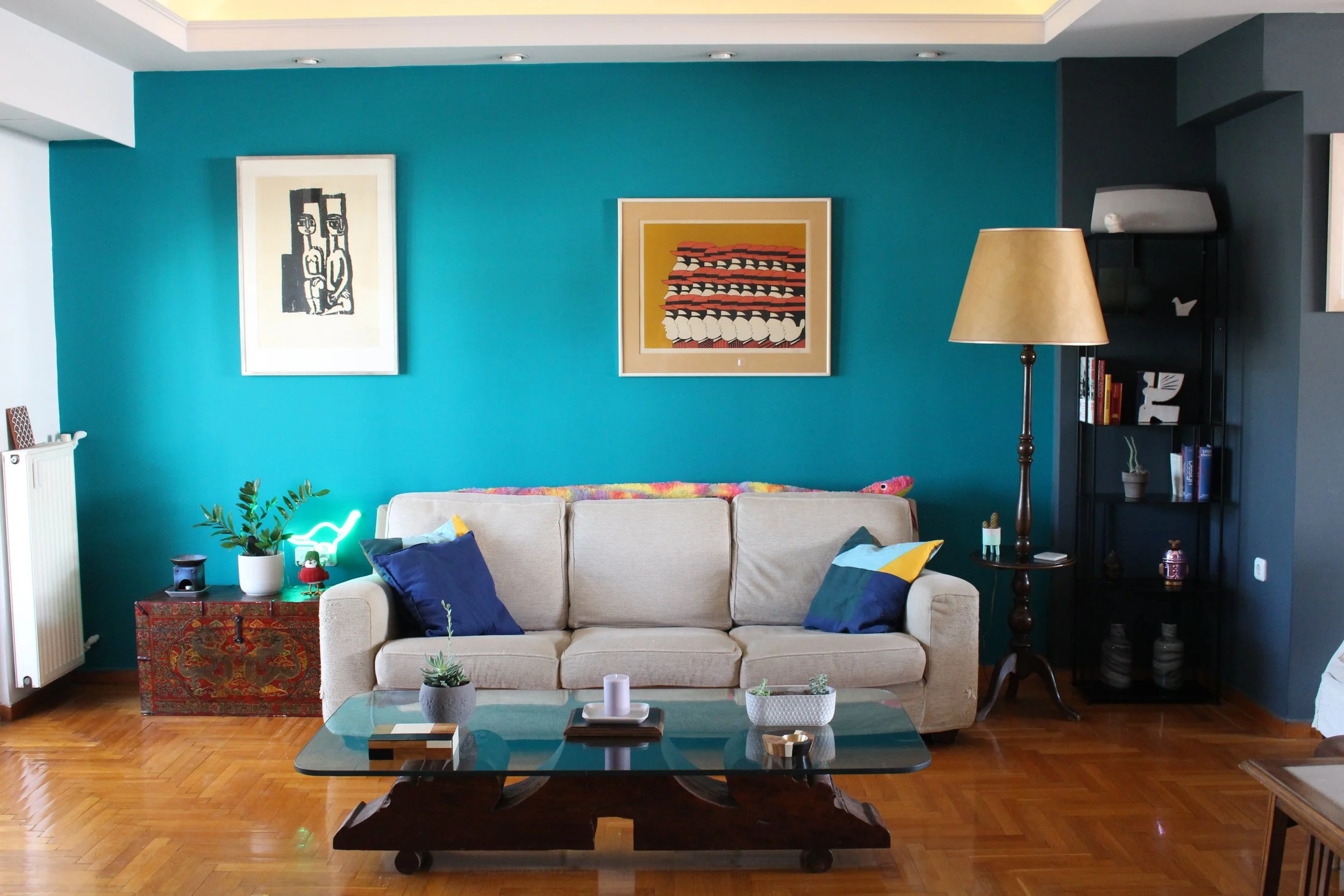 Living room with turquoise accent wall, beige sofa with dark blue and yellow pillows, glass coffee table, colorful paintings, side table with plants and decorative items, black bookshelf with books and decor, wooden floor.