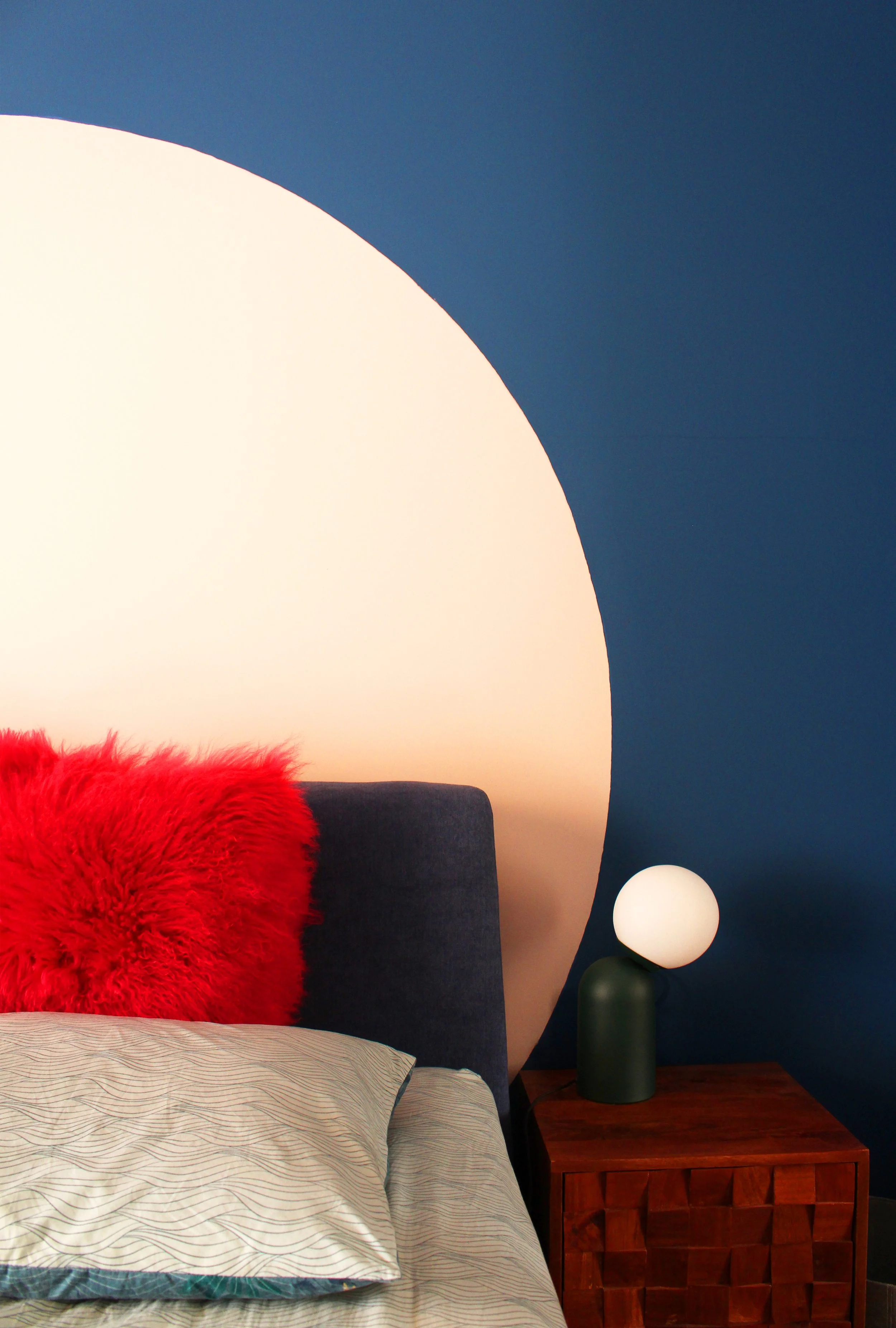 A bedroom with a navy blue wall with a large pink circle to highlight the headboard of the bed, a modern nightstand, a table lamp.