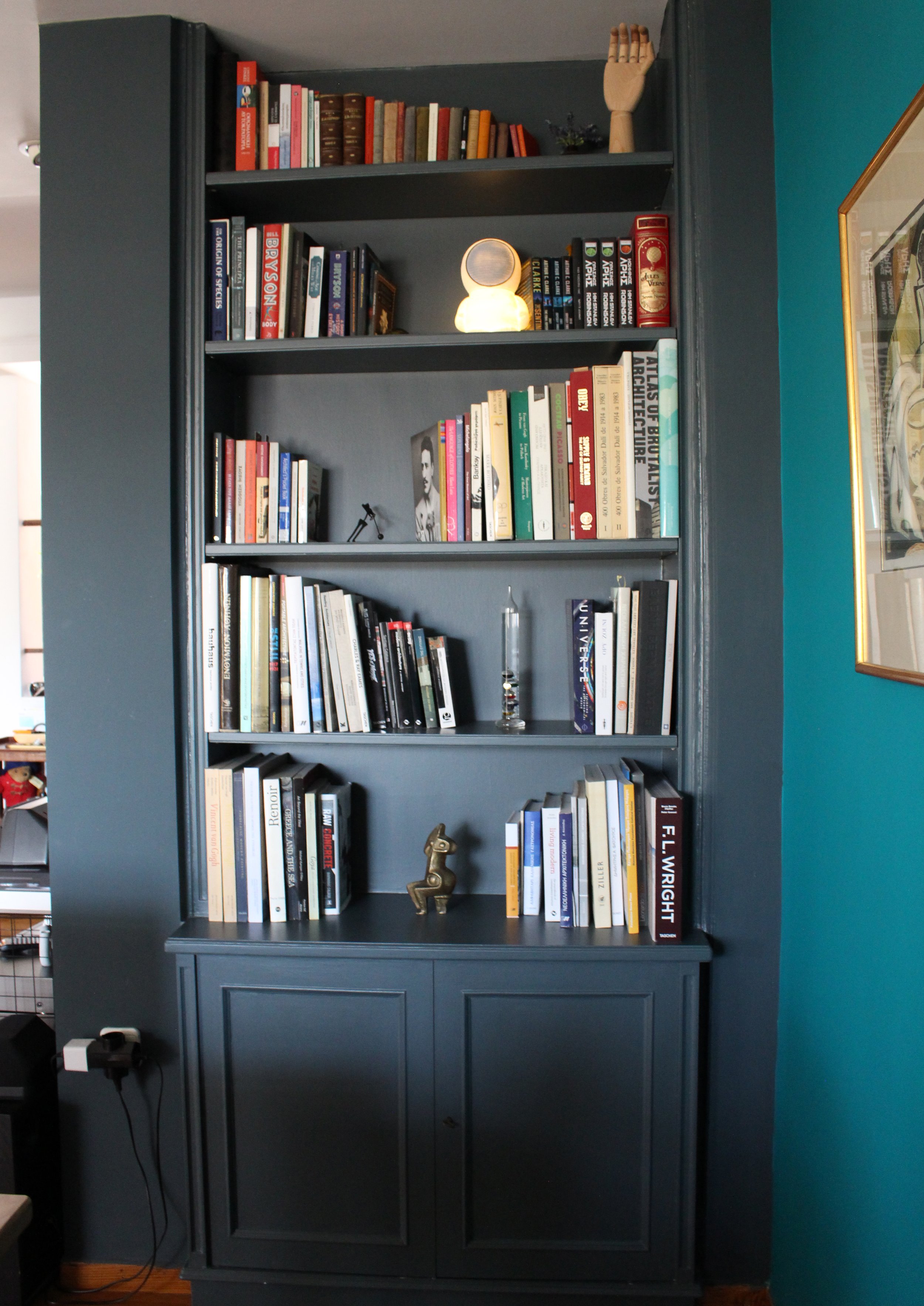 A dark blue bookshelf filled with books, decorative items, a lamp, and a framed picture, set against a teal wall.