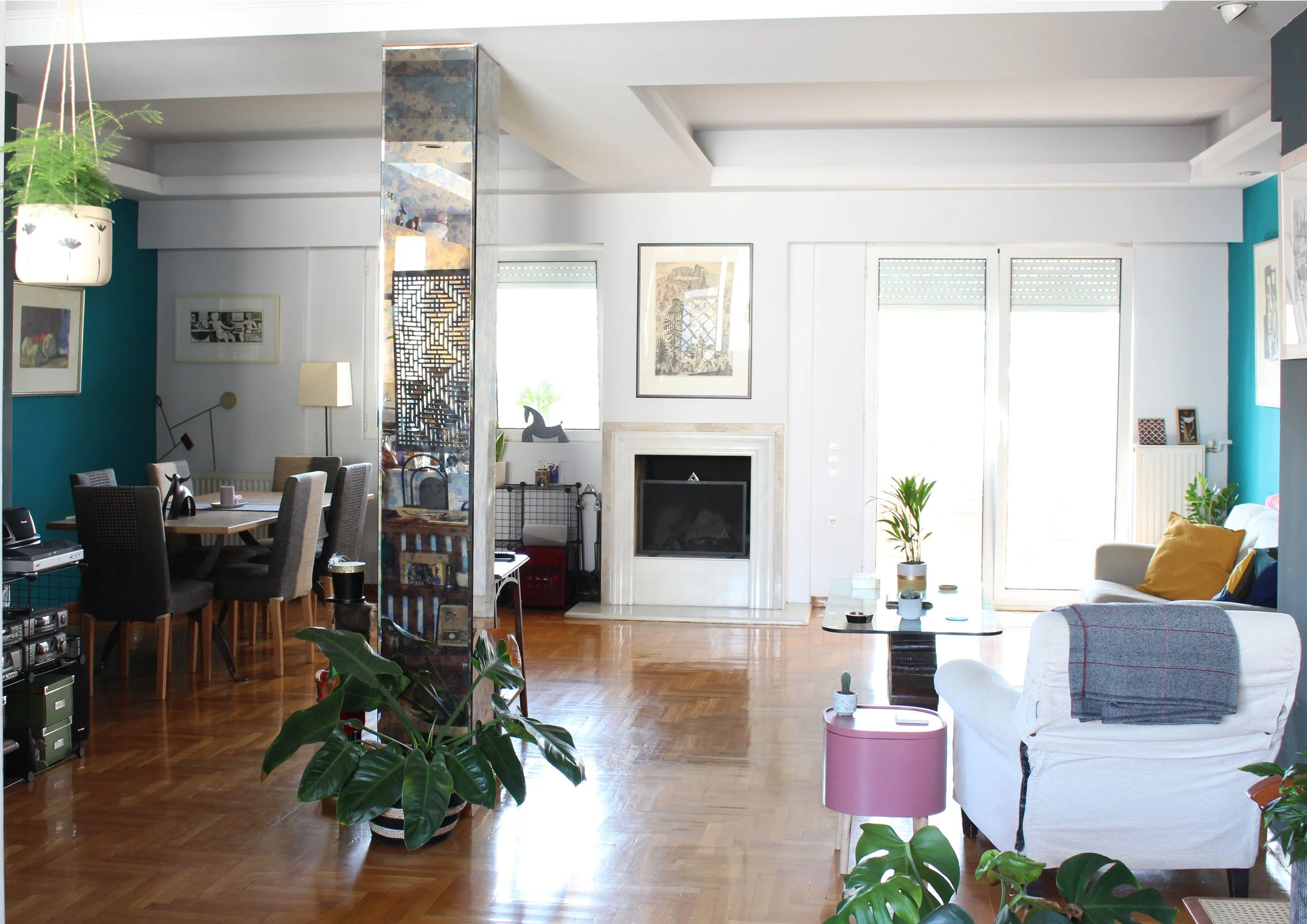 Living room with hardwood floors, white and teal walls, large windows, a fireplace, green potted plants, a white armchair with yellow and blue pillows, a pink side table, a glass coffee table, and a dining area with a table and six chairs.