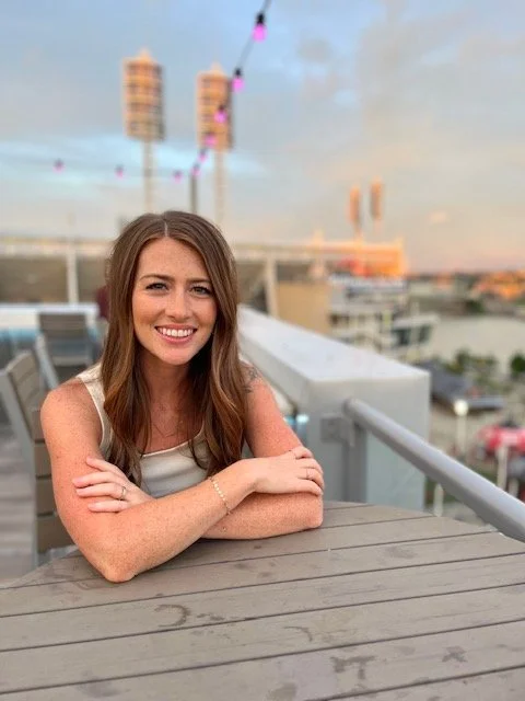 A smiling woman with long brown hair sitting at an outdoor table on a balcony, with a cityscape and a colorful sky in the background.