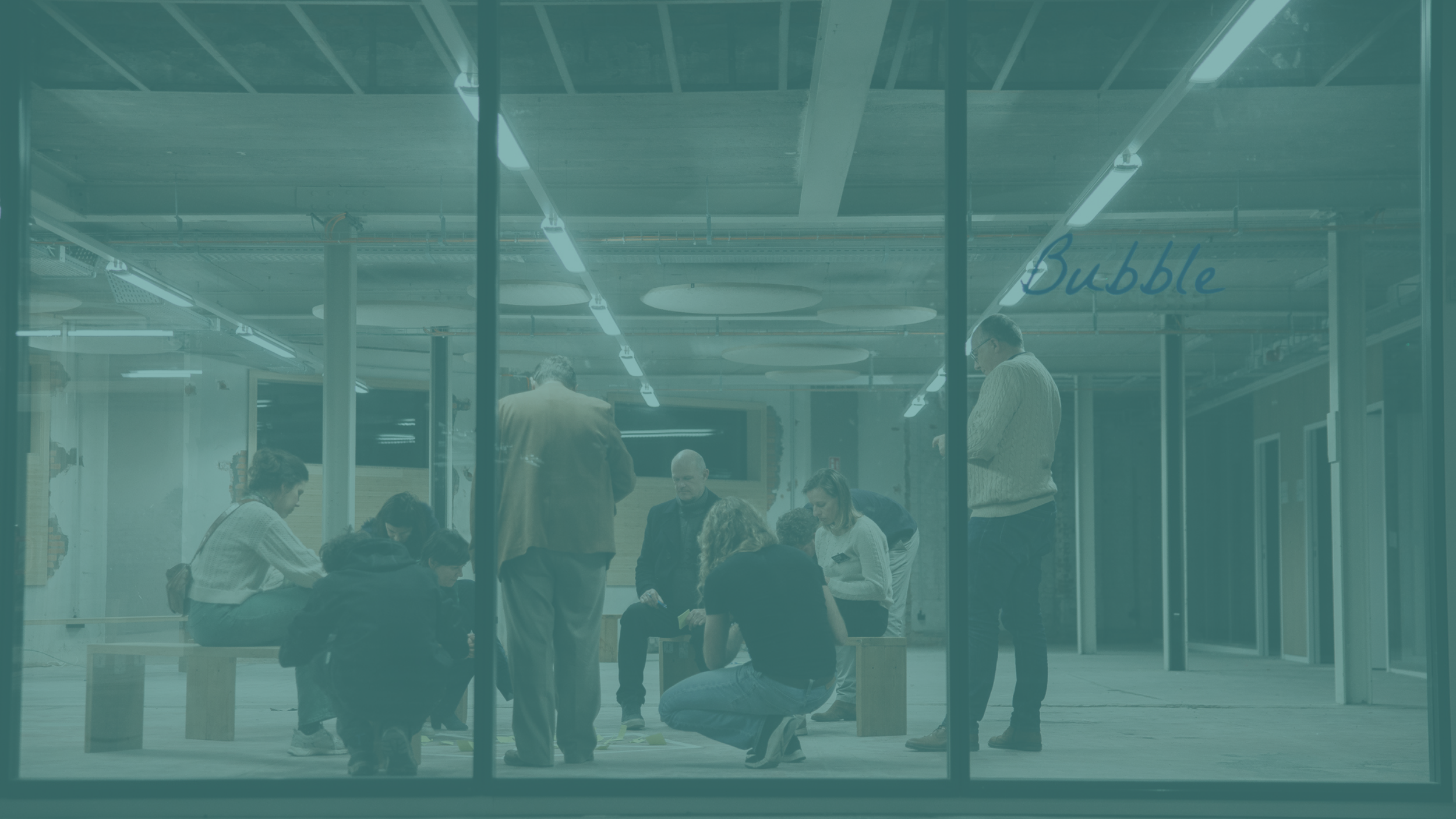 Groep mensen in een kantoorruimte, enkele mensen staan en andere zitten, binnen een moderne omgeving met glazen muren en bureaus, enkele mensen kijken op hun telefoons of laptops.